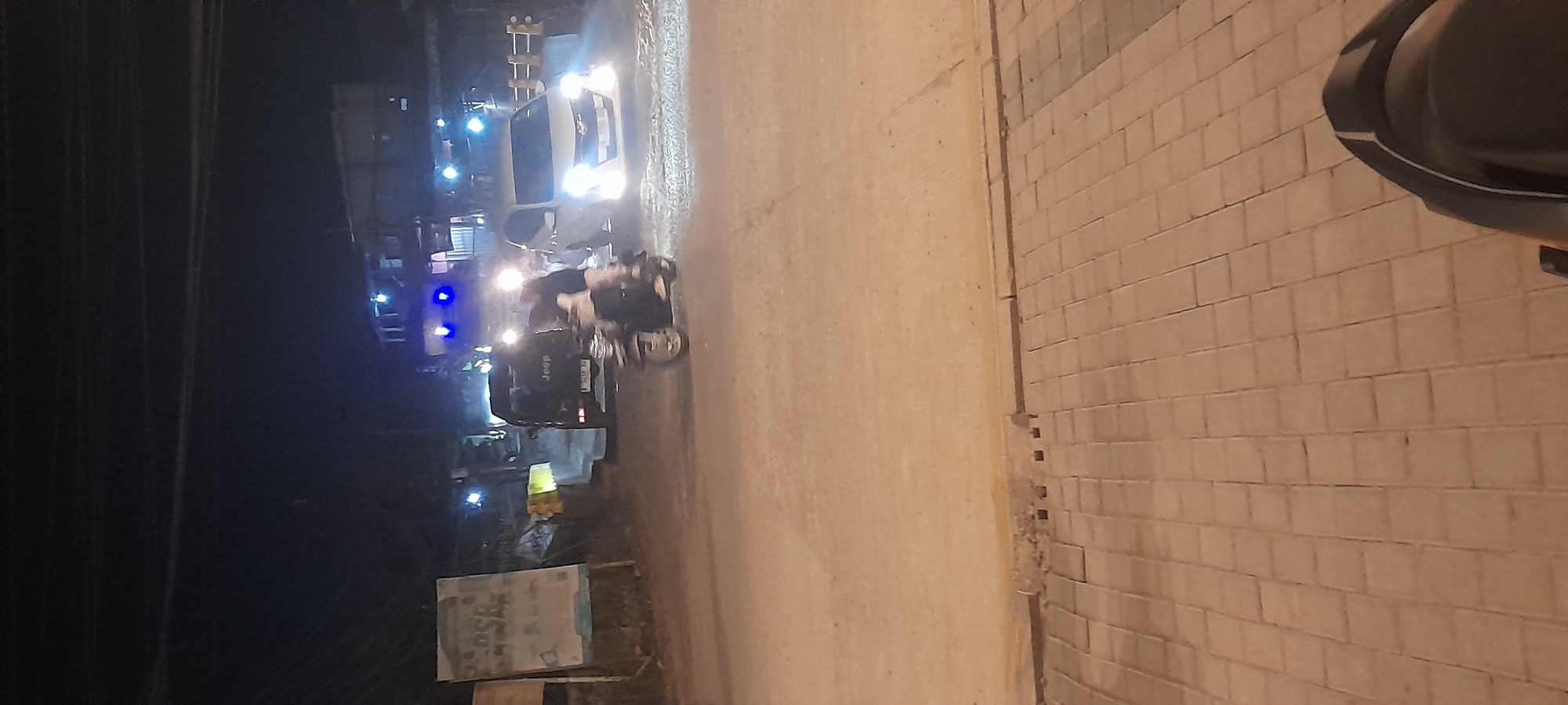 In Kragilan, Indonesia, at night, a motorcycle is ridden on a paved road with a sidewalk lined with interlocking pavers. A black Jeep SUV with the "Jeep" logo visible is stopped on the road. A white sedan with its headlights on is also present. Buildings with illuminated lights are visible in the background. The scene appears to be a street or intersection, with evidence of traffic and urban infrastructure.