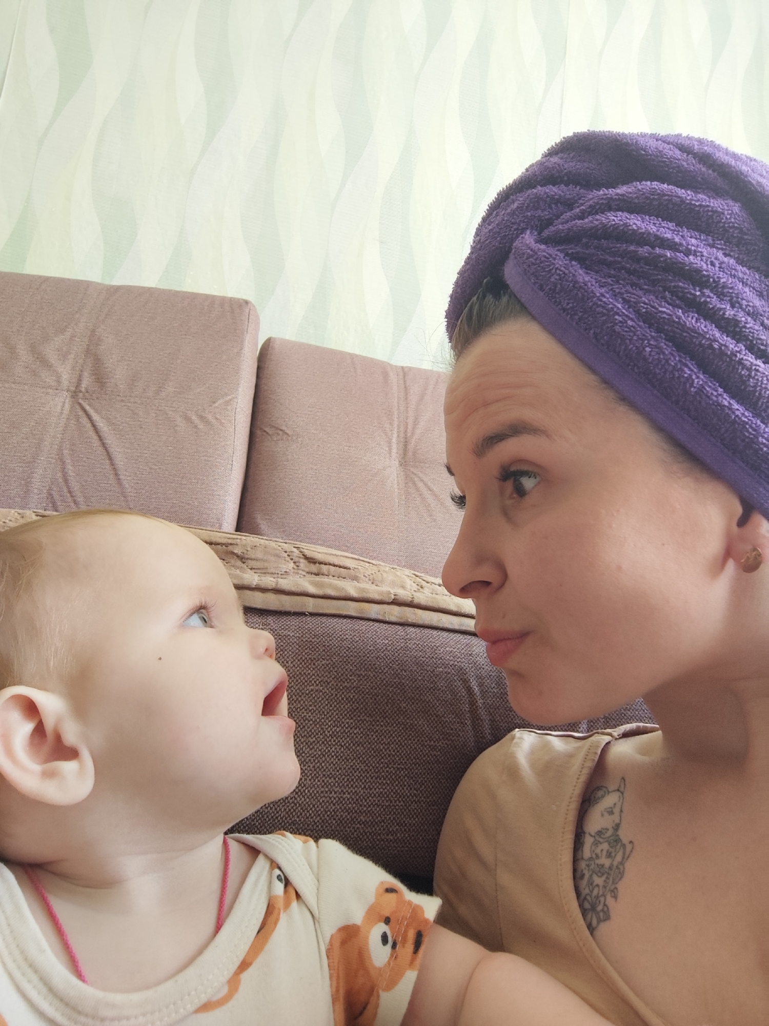 The photograph shows a woman and a baby face to face, seemingly interacting. The woman has a purple towel wrapped around her head. She's wearing a tan-colored shirt and has a tattoo on her chest. The baby is wearing a white onesie with a bear pattern and a pink necklace or string is visible around the baby's neck. A tan-colored couch is partially visible behind the individuals. The background is a light green and white patterned wall. There is no indication this photo was taken in Фоки, Russia.