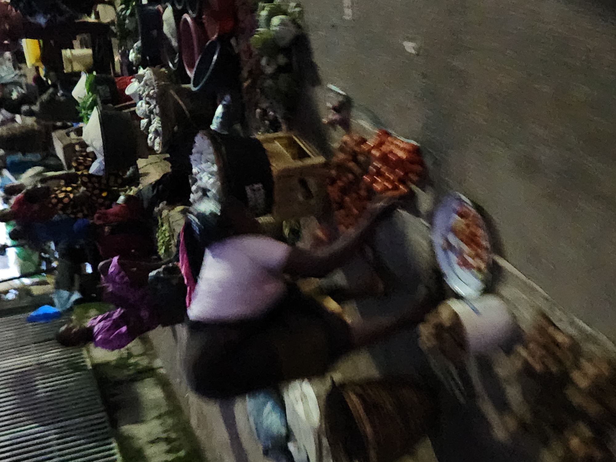 In the city of Kabong, Nigeria, a street market scene unfolds. A person, likely a vendor, is bent over, appearing to sort or display a pile of red items, possibly tomatoes or peppers, arranged on a flat surface. They are wearing a dark skirt and a light-colored top. The surrounding area shows a variety of market activities. In the background, several individuals are visible, some in colorful attire. Various containers, including baskets and buckets, are present, some holding produce like leafy greens and what appear to be white items, possibly onions or garlic. The ground is paved, suggesting an urban setting. The lighting indicates it might be late afternoon or early evening, with some shadows cast by the structures and individuals.