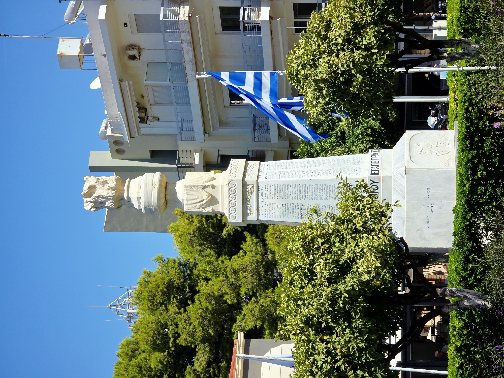 The image depicts a monument in the Agios Nikolaos Municipal Unit, Greece. The monument is a tall, white structure with inscriptions and decorative elements. A Greek flag is prominently displayed, waving in the foreground. Lush green trees and hedges surround the monument, suggesting an outdoor public space. In the background, there are buildings with balconies and air conditioning units, characteristic of a town or city environment. The sky is clear blue, indicating a sunny day. The scene appears to be a memorial or a landmark within a well-maintained urban area. There are no visible people interacting with the monument.