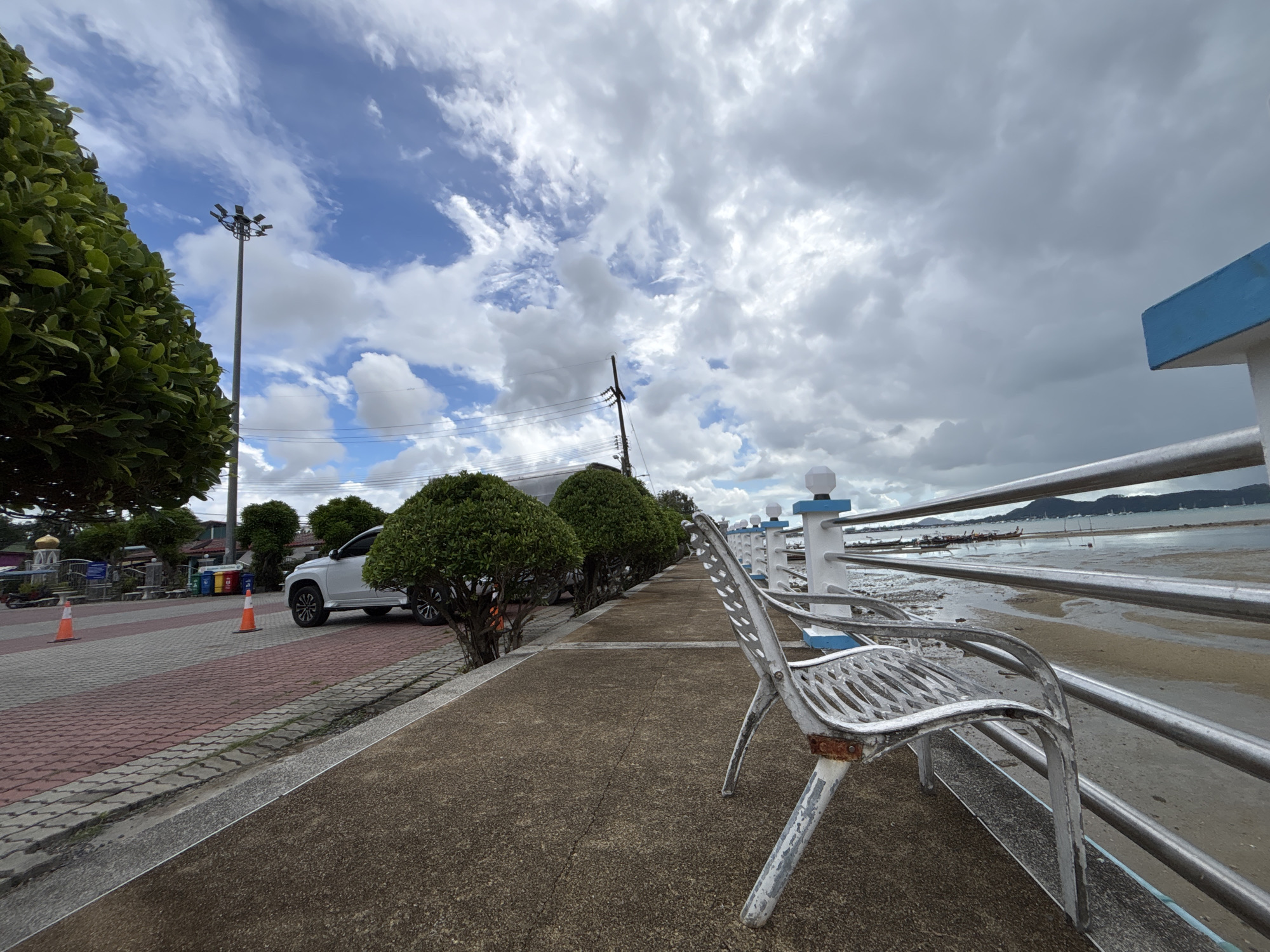 The image depicts a waterfront promenade in Rawai, Thailand. In the foreground, a weathered, white metal bench with rusted accents sits on a concrete pathway. To the left of the bench, a paved walkway with red and grey bricks is visible, lined with manicured, spherical green bushes. Beyond the bushes, a white SUV is parked. A tall street lamp with multiple lights stands near the bushes. In the background, a silver metal railing with blue accents separates the promenade from the water. The water is shallow, revealing a muddy seabed, with several long-tail boats anchored offshore. In the distance, a hazy coastline with mountains can be seen under a cloudy sky. The overall impression is of a public space near the sea, likely a scenic spot in Rawai.