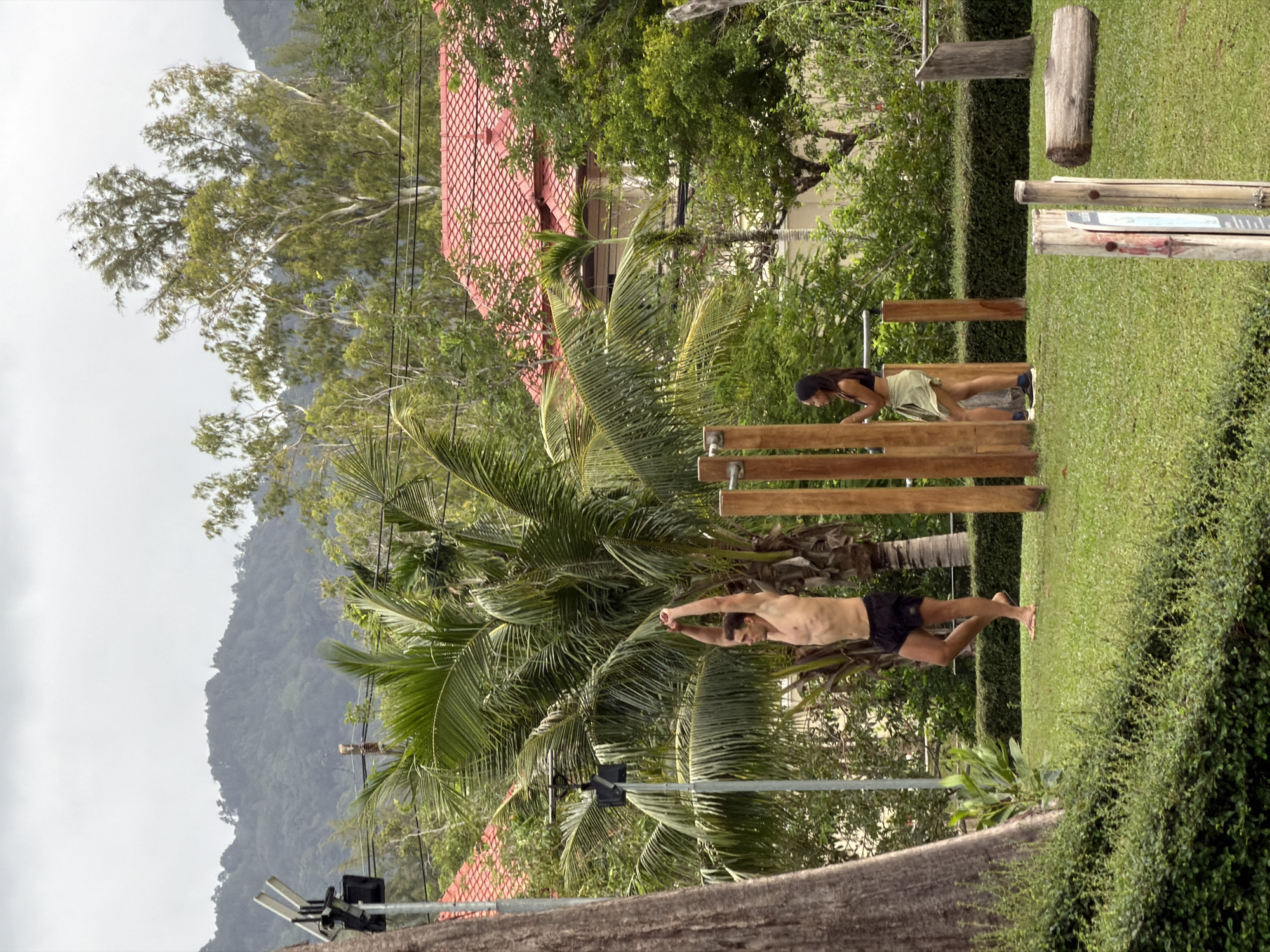 The image depicts an outdoor gym area in Choeng Thale, Thailand, situated on a grassy field with lush vegetation and palm trees. In the foreground, a man is stretching with his arms raised overhead, wearing black shorts. Nearby, a woman stands by a wooden pull-up bar, wearing a light-colored skirt and a dark top. Behind them, a residential building with a red tiled roof is visible, surrounded by dense greenery and hills in the distance. The sky is overcast.