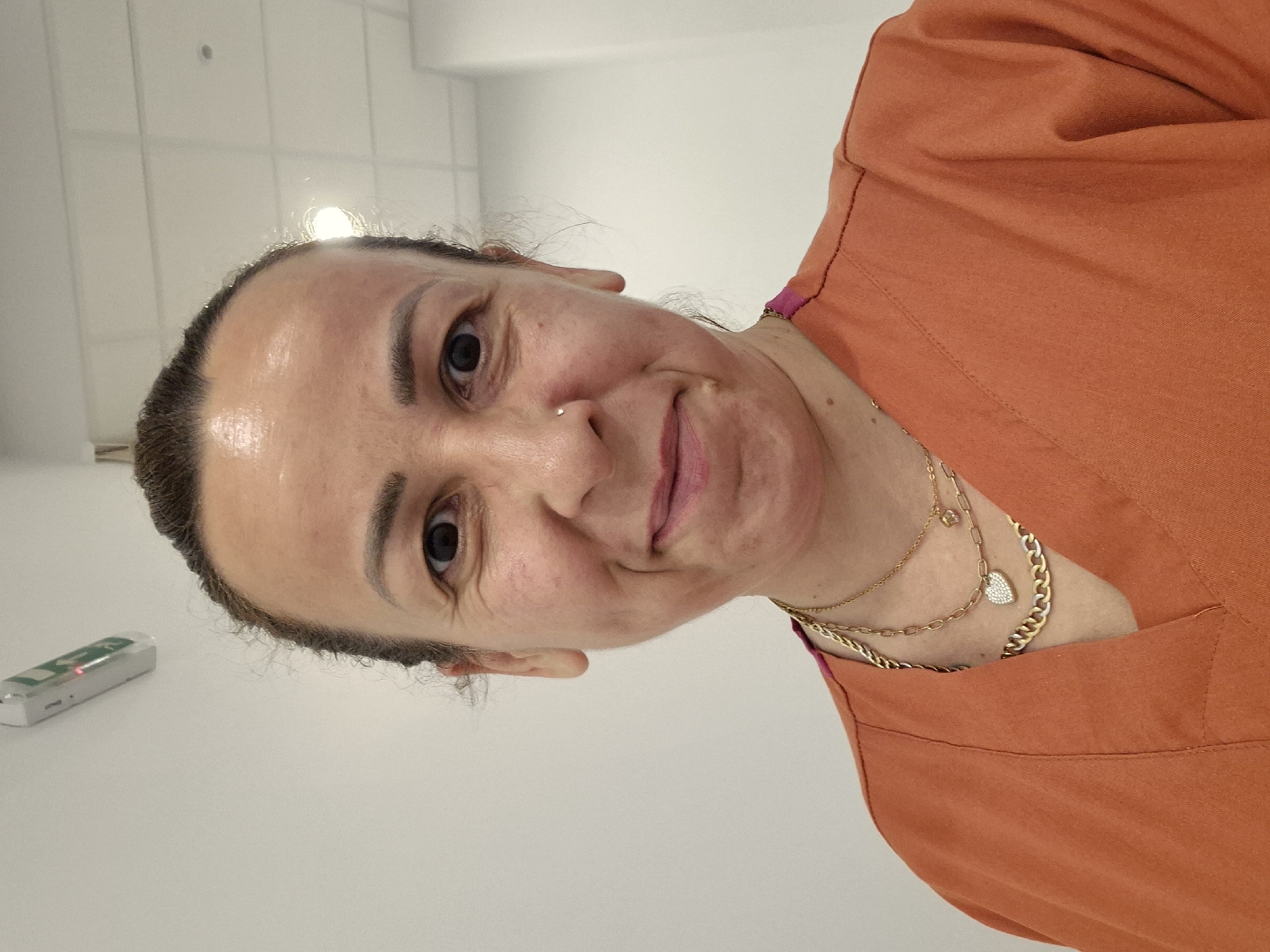 A medium shot photograph shows a woman with medium-brown hair pulled back from her face, looking directly at the camera with a slight smile. She is wearing an orange scrub top with a pink trim at the neckline. She has a nose ring and is wearing multiple gold necklaces. One necklace has a heart-shaped pendant. The background is a plain white wall with a light fixture on the ceiling. The overall impression is that the photo was taken indoors in a clinical or professional setting.