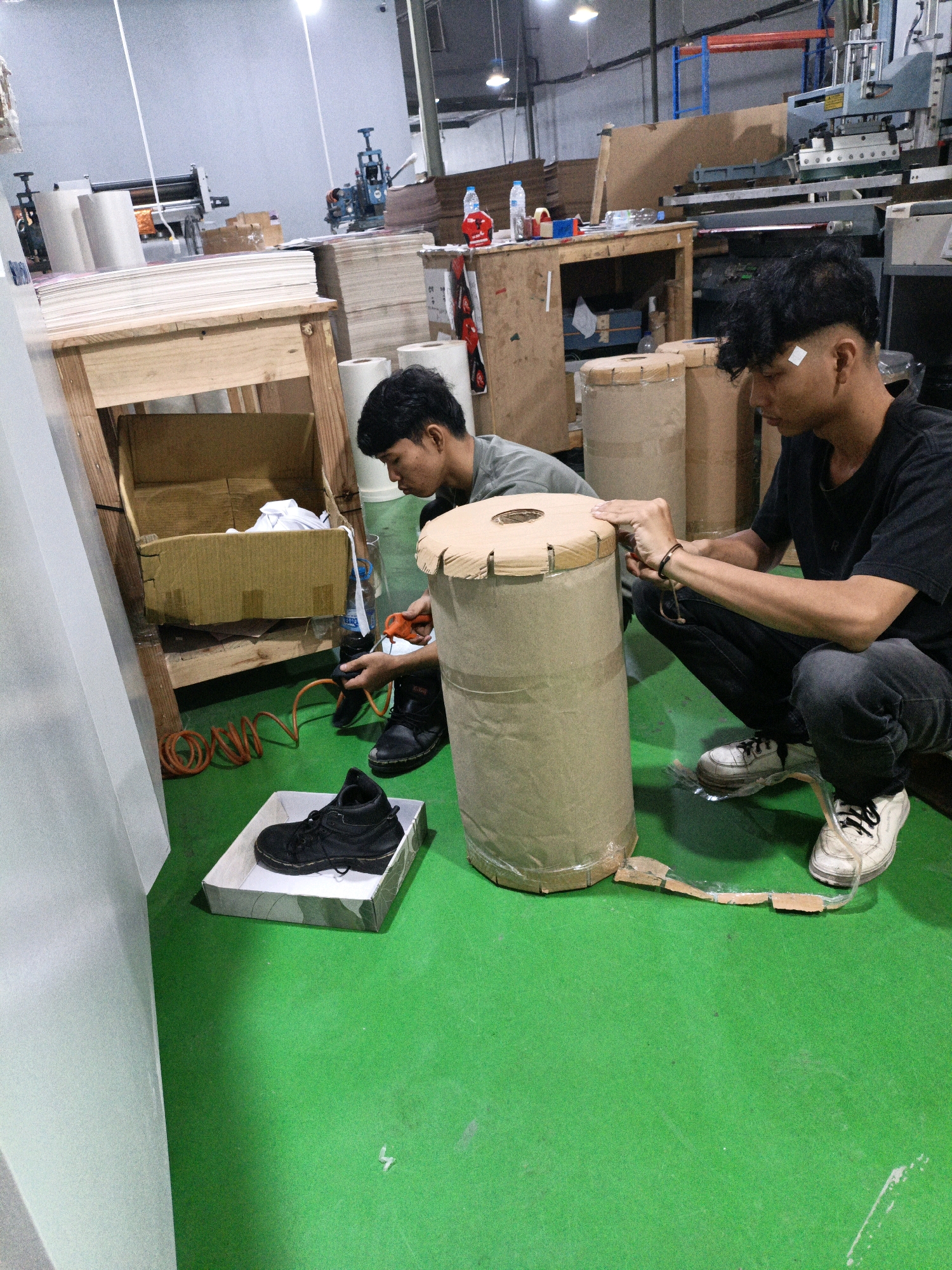 In a factory setting in North Jakarta, Indonesia, two young men are engaged in work. The floor is a vibrant green, and industrial machinery and materials are visible in the background. In the foreground, a large cardboard cylinder, wrapped in brown paper and tape, stands upright. One man, wearing a black t-shirt and black jeans, is squatting and appears to be working on the cylinder. He is wearing white sneakers. Another man, wearing a grey t-shirt, is partially visible in the background, also appearing to be involved in the task. A black work boot sits in a white and camouflage patterned box on the floor. An orange coiled hose and an orange handheld tool, possibly an air gun, are also present on the floor. The overall scene suggests a process of packaging or preparation of materials within the factory.