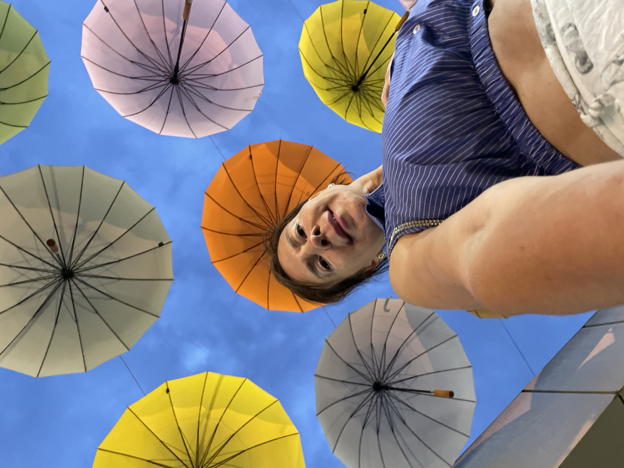 A woman is looking up at the camera, surrounded by colorful umbrellas suspended in the sky. She is wearing a blue and white striped shirt and white pants. The umbrellas are yellow, orange, pink, and white, and they are arranged in a pattern against a blue sky. The image appears to have been taken from a low angle, looking up at the woman and the umbrellas. The location is stated to be Sochi, Russia, but there are no specific visual cues to confirm this.