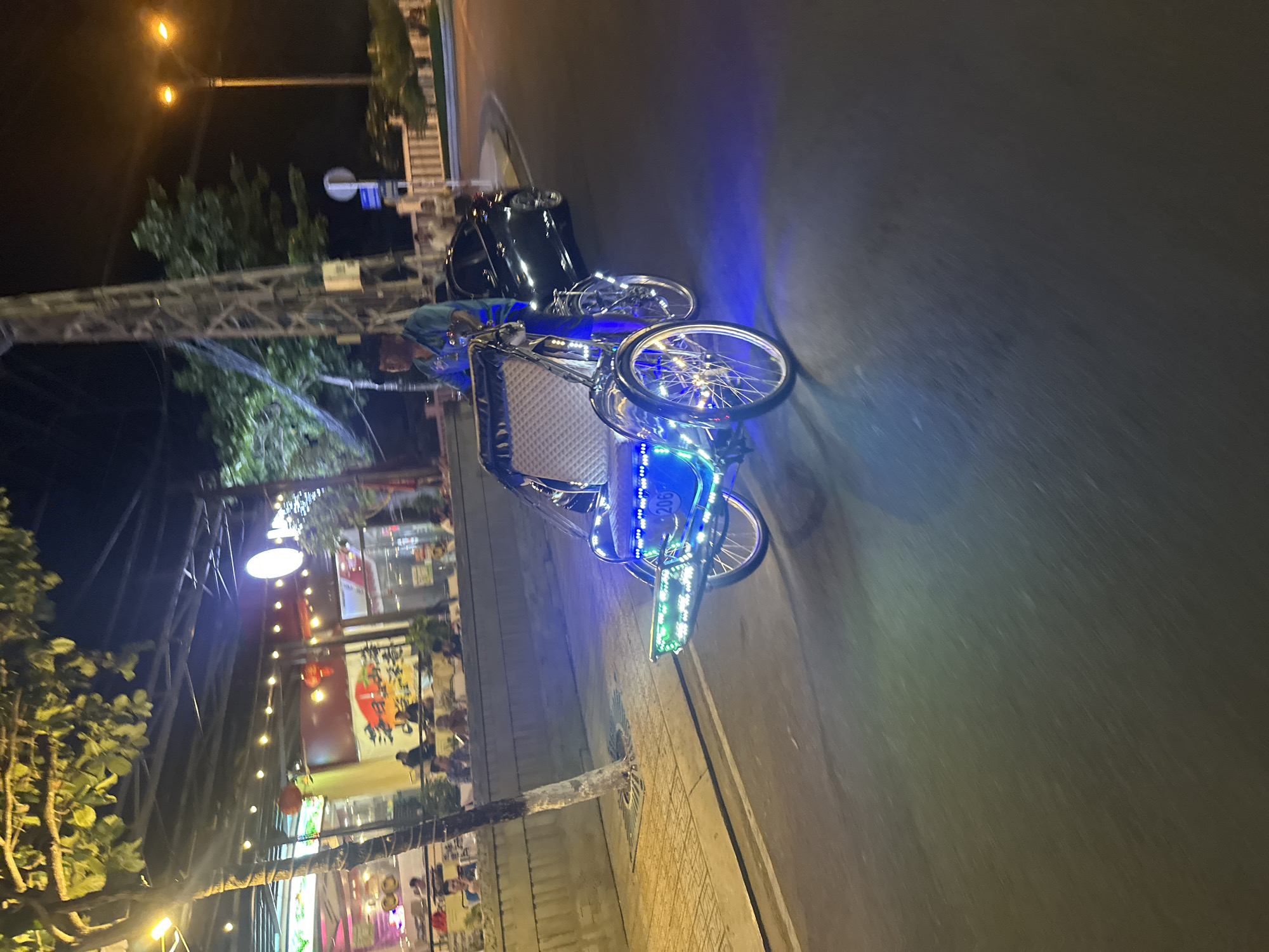 The image captures a nighttime street scene in Bac Nha Trang Ward, Vietnam. A brightly lit cyclo (a three-wheeled passenger vehicle) stands on the sidewalk, adorned with blue and green LED lights, possibly indicating its availability for hire. The cyclo's seat is visible, as are its wheels and frame. In the background, a restaurant or cafe is open, with patrons visible inside. String lights and other decorative lighting illuminate the building's exterior. The street itself appears dark, with a car parked nearby. Streetlights are present, casting a glow on the surroundings. The perspective is slightly tilted, adding a dynamic feel to the scene.