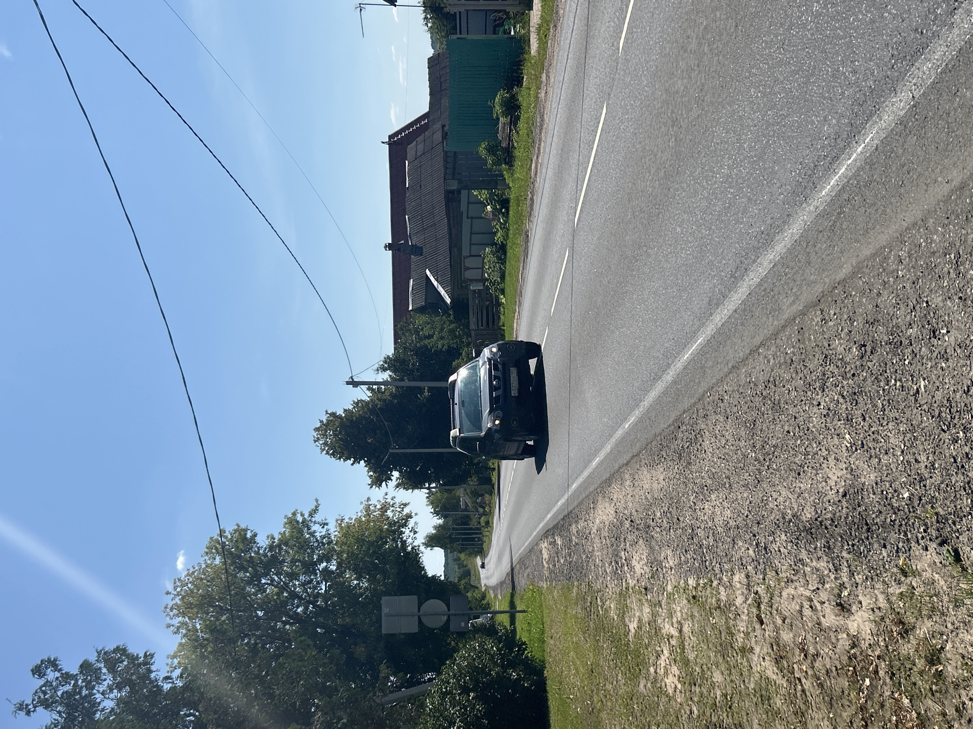A dark-colored SUV is driving on a paved road in an outdoor rural setting. The road has a white dashed line down the center and a solid white line on the right edge. The car is in the right lane, moving away from the camera. The driver's side is visible, and the license plate is partially visible. To the left of the road is a gravel shoulder with some grass and bushes. Beyond the shoulder is a grassy embankment and trees. To the right of the road, in the background, are several houses with dark roofs, a green fence, and more trees. The sky is blue with a thin white contrail. Power lines are visible stretching across the sky. The scene appears to be during the daytime under clear skies. The location is identified as Kalitsino, Russia.