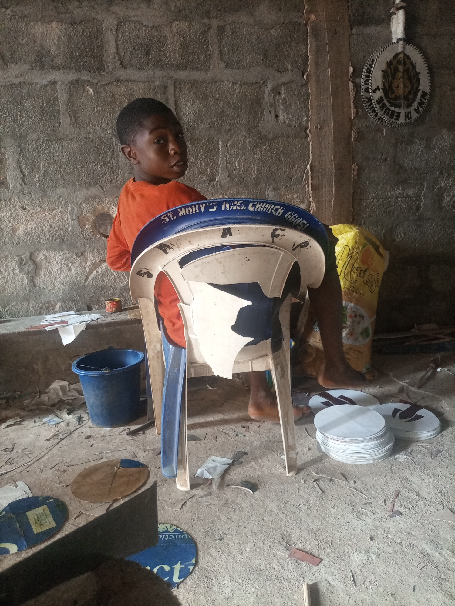 This media file captures a young Black boy sitting on a plastic chair in what appears to be an indoor workshop or storage area. The boy is wearing an orange t-shirt and is looking over his shoulder towards the camera with a neutral expression. He is seated on a blue and white plastic chair that has "ST. MARY'S ANG. CHURCH GROSI" written on its back.
The setting is rustic, with a rough brick wall in the background and a dirt or concrete floor scattered with debris, paper scraps, and some circular objects. To the right of the boy, a large yellow bag, possibly containing rice, is leaning against the wall. On the floor in front of him, there is a stack of white circular objects, some of which have dark brown or red markings. Other loose circular items are scattered around the floor, including one that appears to be a flattened kite with blue and brown segments. A blue bucket is also visible on the floor to the left.
On the wall behind the boy, a circular decorative item with text and an image is mounted. Part of the text visible on this item appears to be "NINA DI EBUBE".
The lighting suggests an indoor setting, possibly during the daytime, with natural light filtering in from an unseen source. The overall impression is one of a utilitarian workspace, possibly related to craft or kite making.