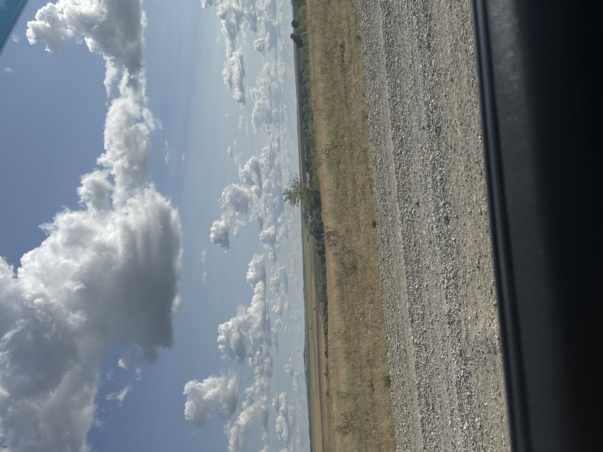 The image depicts a rural landscape under a cloudy sky. In the foreground, there is a gravel road. The middle ground consists of a dry, grassy field with some scattered bushes and a small tree. In the background, there are rolling hills with patches of tilled land and more vegetation. The sky is blue with large, fluffy white clouds. The scene is devoid of people or any visible human activity. It is unlikely that this image is of Васильевка, Russia, as the landscape does not appear to be urban or suburban.