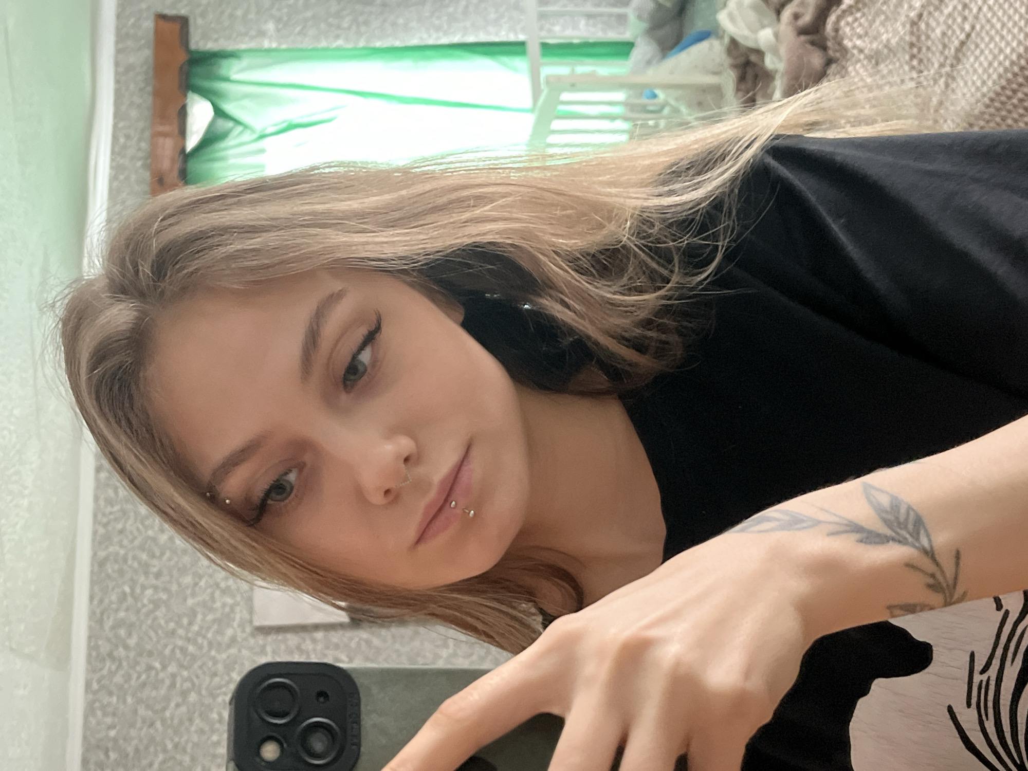 A young woman with blonde hair and facial piercings, including one on her eyebrow, nose, and lower lip, is taking a selfie. She is wearing a black shirt and has a tattoo on her left wrist. She is holding a smartphone with a triple-lens camera. The background includes a window with a green covering, a white chair, and a bed with patterned bedding. The location is not discernible from the image.