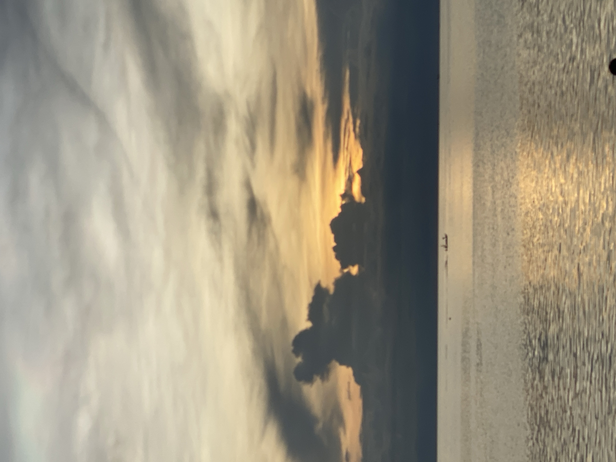 This media file captures a serene sunset over the ocean, likely in Koh Phangan, Thailand. The scene is dominated by dramatic clouds painted in hues of orange, yellow, and gray as the sun dips below the horizon. The water reflects the sky's warm tones, creating a shimmering, textured surface.
A small, dark silhouette of what appears to be a boat or a very small vessel is visible on the water, adding a sense of scale and tranquility to the vast expanse. The overall mood is peaceful and contemplative, evoking the beauty of a tropical evening. The time of day is clearly sunset. There is no visible text in the image.