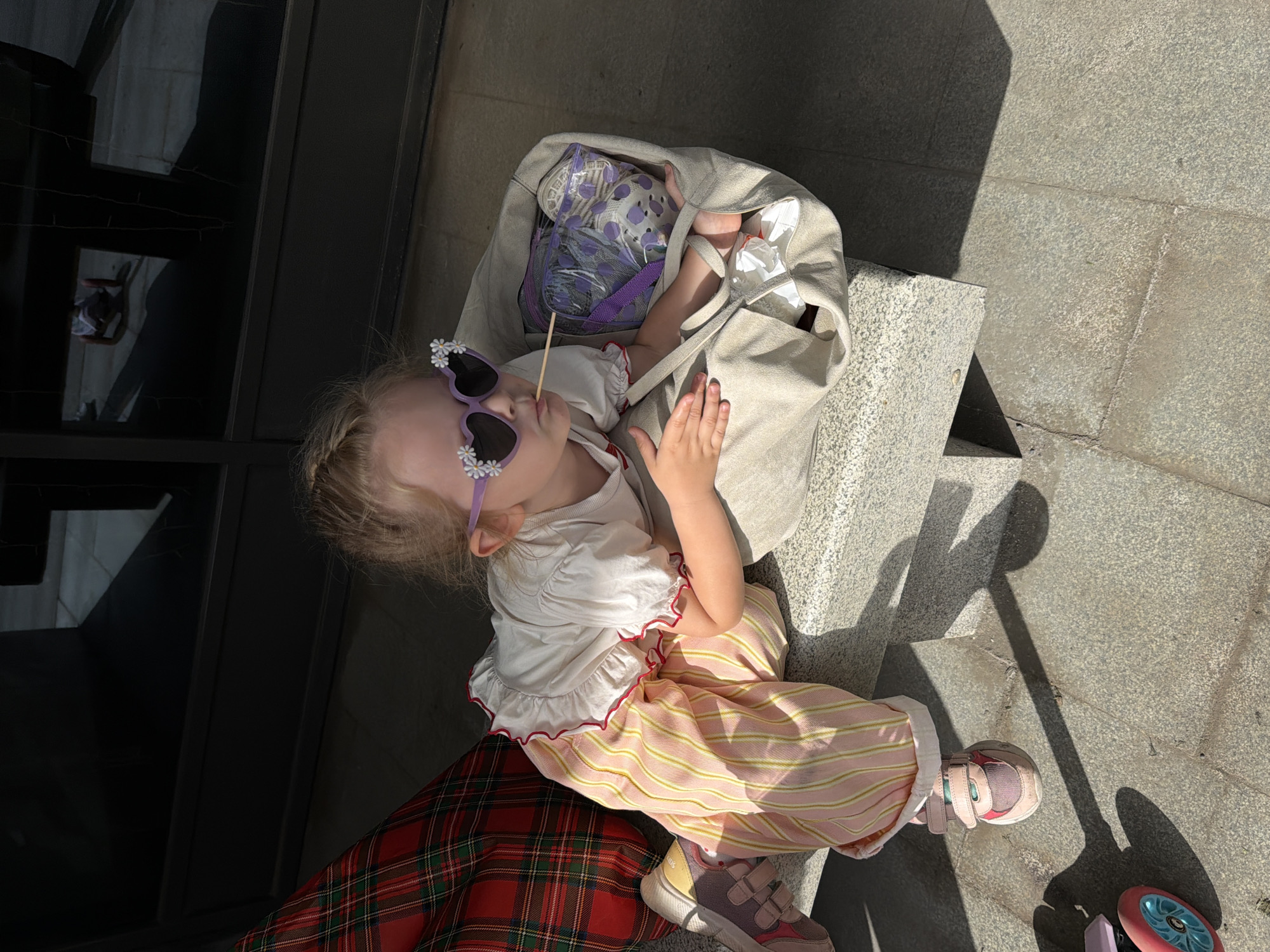 A young child sits on a low concrete ledge outdoors in Saratov, Russia. The child wears heart-shaped sunglasses decorated with daisies, a white top with red trim, pink and yellow striped pants, and pink sneakers. They are sucking on a wooden stick. A large beige bag sits next to the child, containing various items including a purple mesh bag with polka dots. The ledge sits against a dark glass wall on one side and a concrete walkway on the other. Sunlight casts shadows on the walkway. Part of a red plaid blanket or garment is visible beneath the child. A scooter and its shadow are visible near the child's feet.