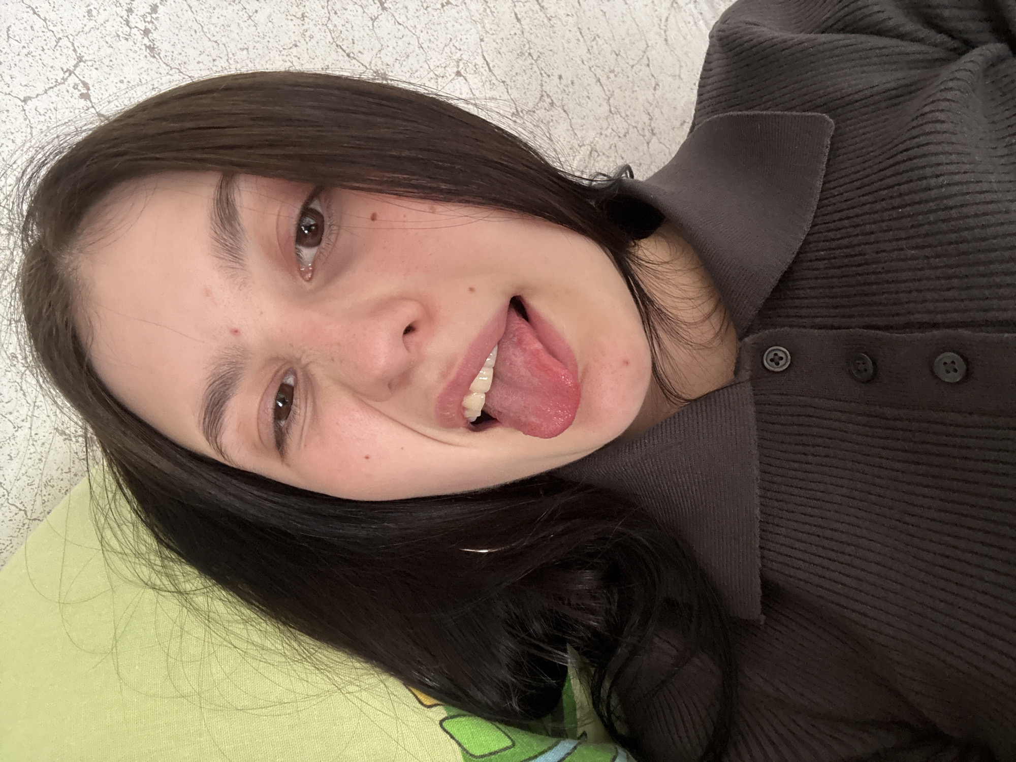 A young woman with long, dark hair is lying down, possibly on a pillow with a light green patterned cover. She is wearing a dark gray collared shirt with buttons. She is sticking her tongue out and making a face. The background is a textured, light gray wall. There is no indication of the location being in Фоки, Russia.