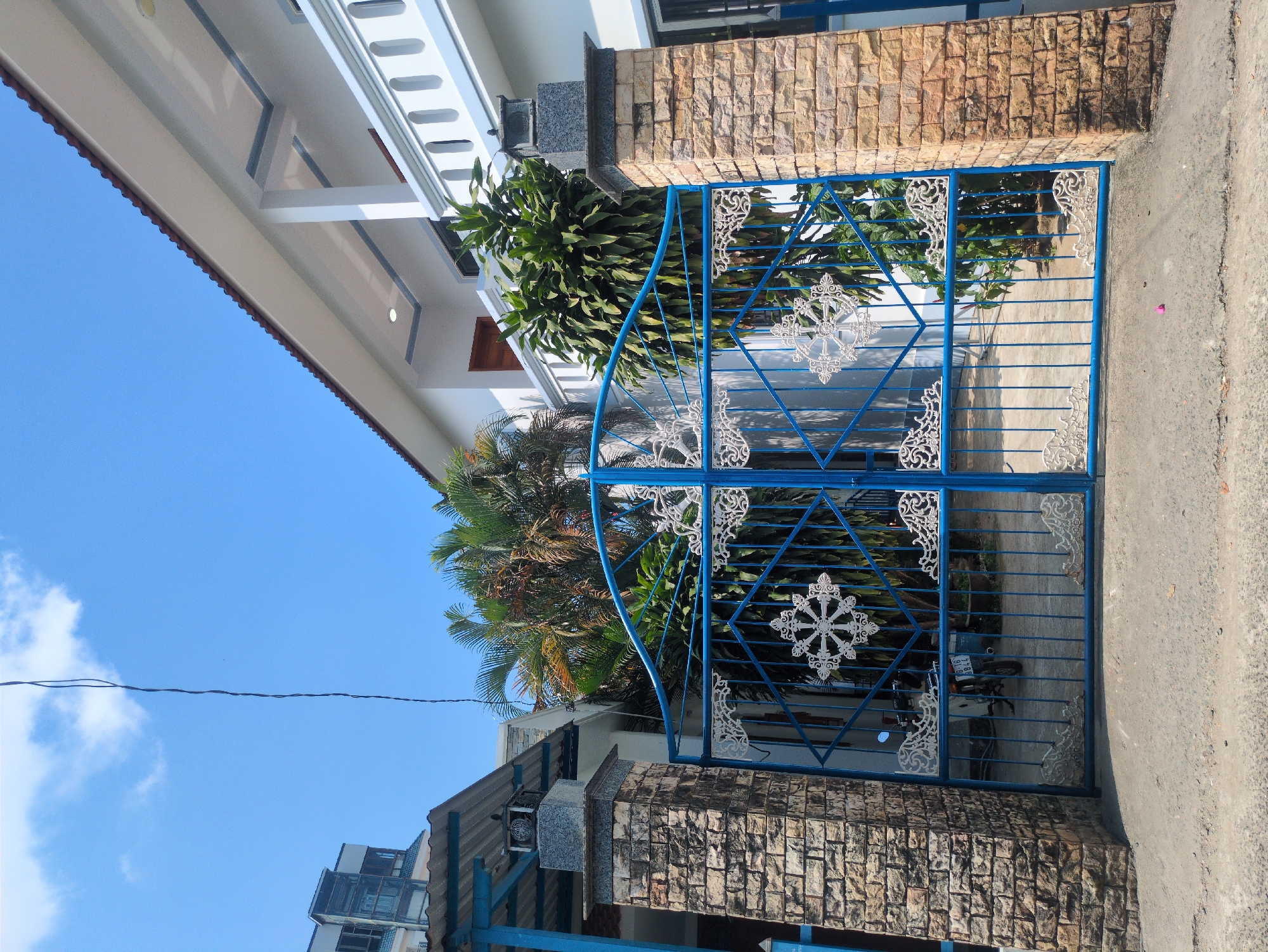 The image shows the exterior of a building in Phú Quốc, Vietnam. A blue ornate metal gate with white decorative accents is the central element. The gate is set between two pillars constructed of rough-hewn light brown stones. Behind the gate, partially obscured by the gate's design and some foliage, a parked motorcycle is visible. The gate is situated on a concrete pathway. To the left of the gate, part of a building with a corrugated metal roof can be seen, with a taller building in the background. Above the gate, a palm tree and other greenery are visible. To the right of the gate, a two-story building with white trim can be seen, and part of the bright blue sky is in the background. A thin electrical cable is visible in the sky, spanning the top of the image.