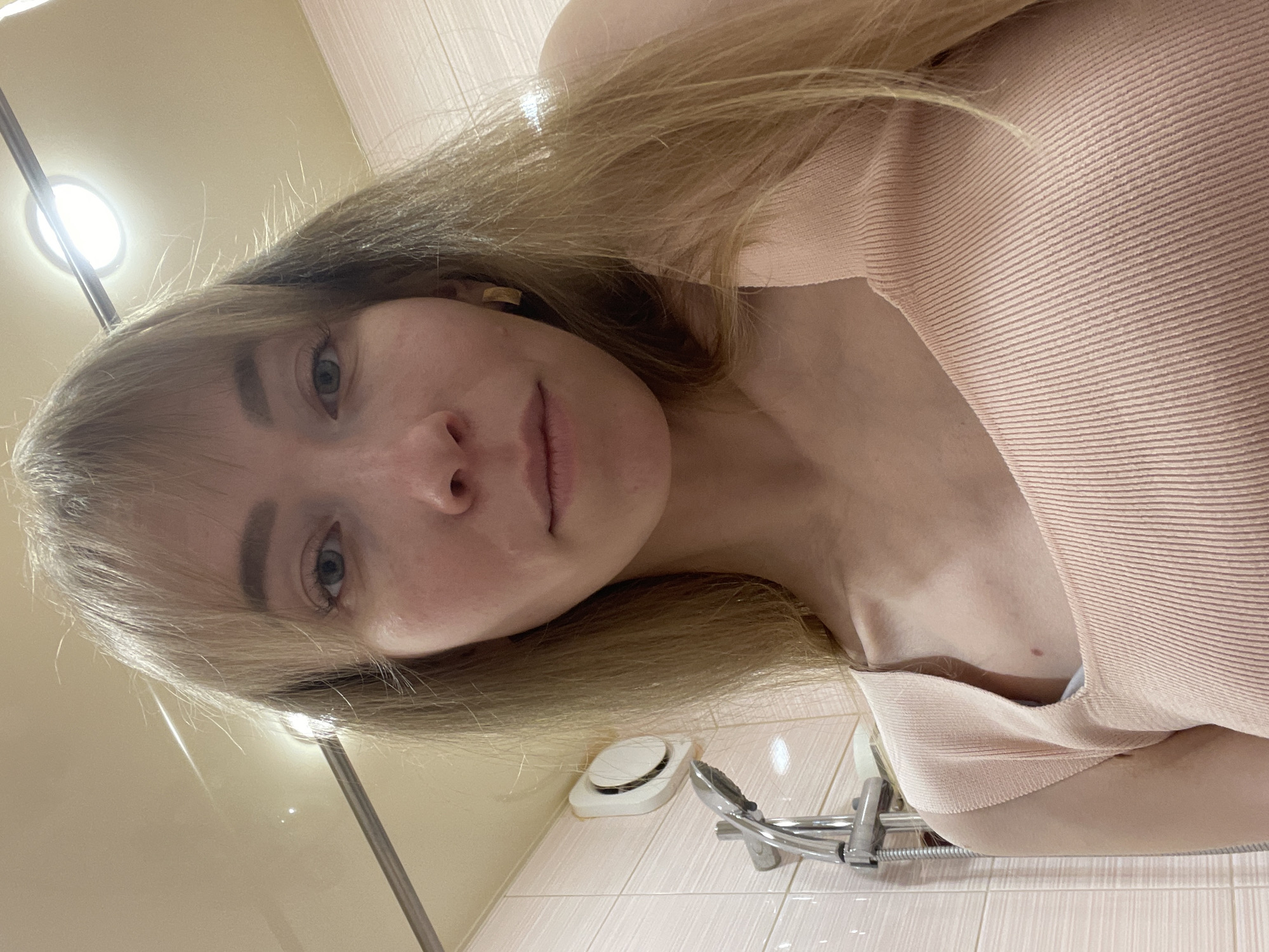 A woman with blonde hair and fair skin is taking a selfie in a bathroom. She is wearing a light pink ribbed top. The bathroom has tiled walls and a showerhead. A vent is visible on the wall. The location is presumed to be Perm, Russia.