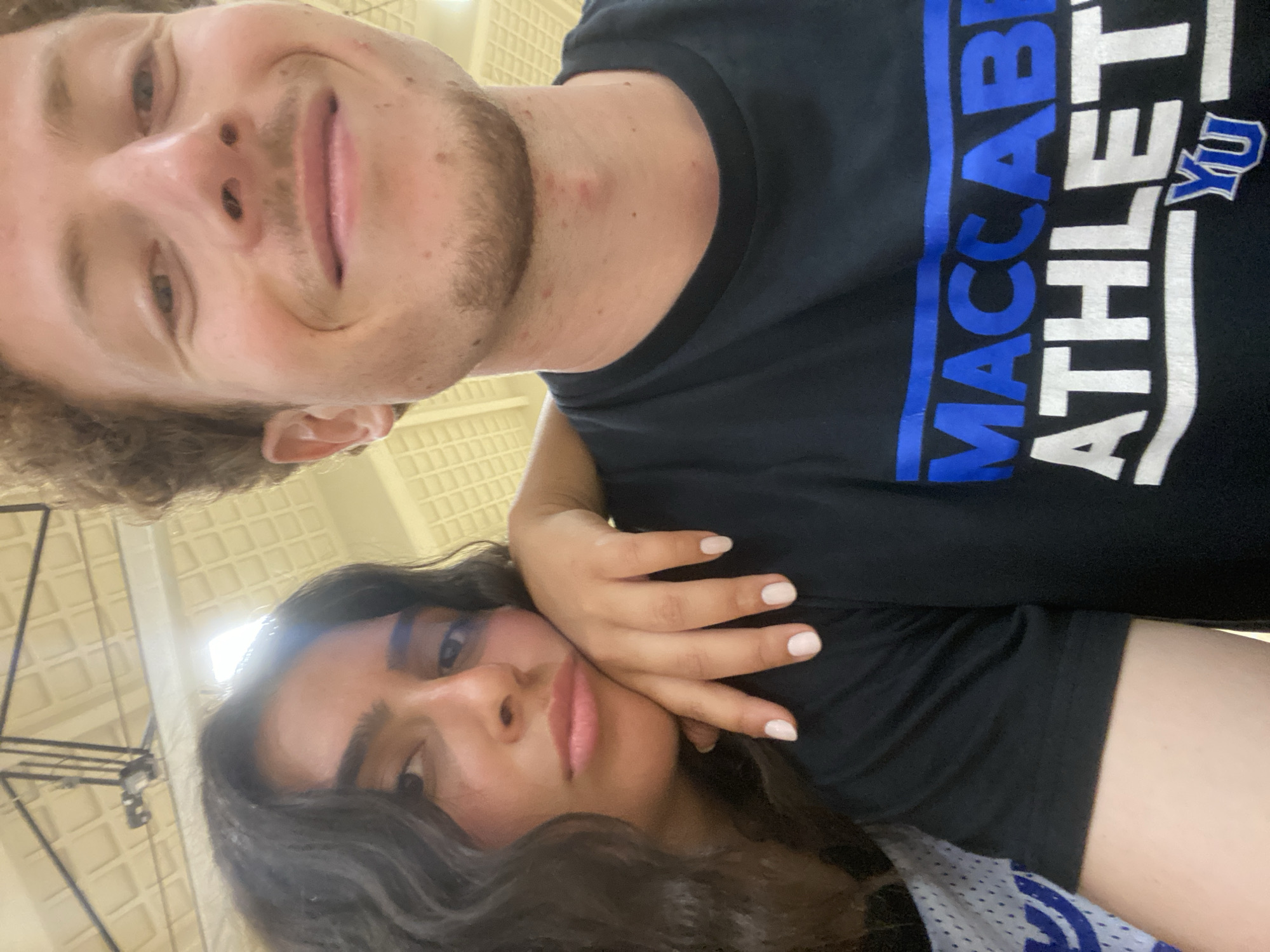 A selfie image depicts two individuals in an indoor setting, likely a gymnasium, given the overhead lighting and ceiling panels. The male, situated at the top of the frame, has fair skin, curly hair, and facial stubble. He is wearing a black T-shirt with the words "MACCABI ATHLETE" in blue and white lettering along the shirt. The female, positioned at the bottom of the frame, has dark hair and fair skin. Her hand is placed near her face. The image appears to have been taken at the University of Maryland's College Park campus.