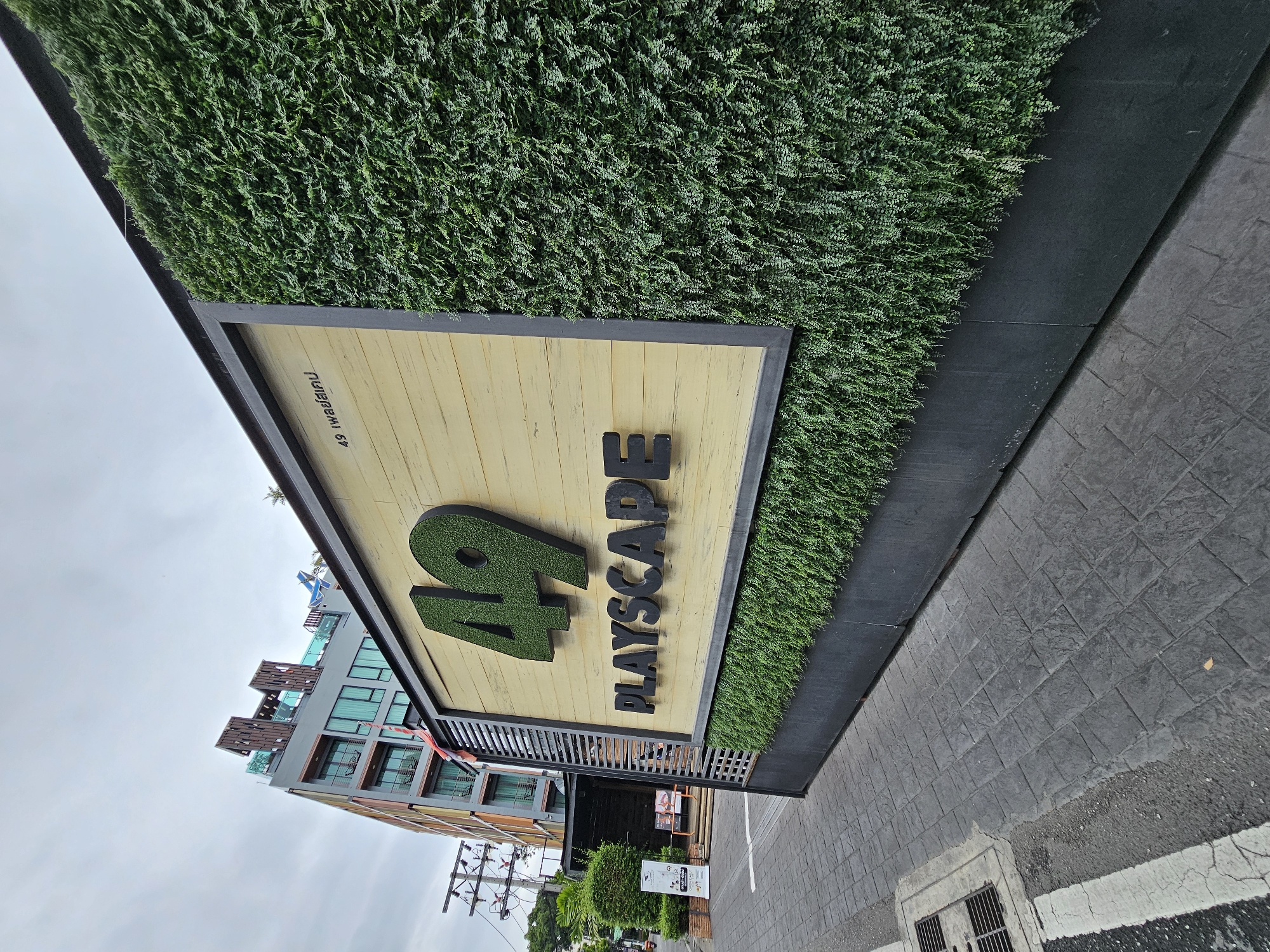 The image shows a building facade at the entrance to "49 PLAYSCAPE" in Bangkok, Thailand. The facade features a light-colored wooden panel with a large green "49" logo and the text "PLAYSCAPE" in black. The logo and text appear to be three-dimensional. The right side of the facade is covered in a dense, artificial green plant wall. To the left of the entrance, a modern building with multiple stories and balconies is visible. The foreground shows a paved sidewalk with a gray, patterned surface and a metal grate on the left. The sky is overcast and gray. There are no people clearly visible in the image, though there are some indistinct figures in the background near the building entrance. A Thai flag is attached to the left side of the entrance structure.
