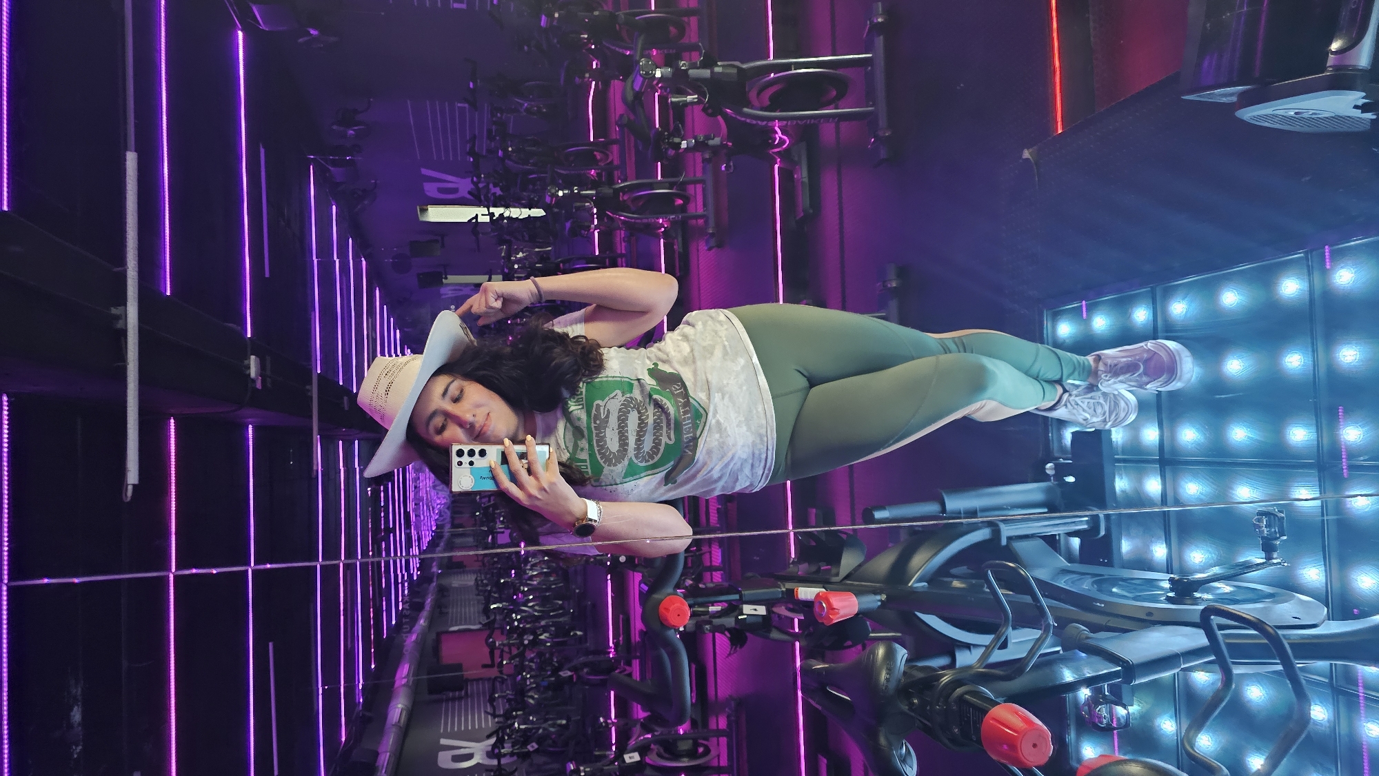 A woman is taking a selfie in a mirror. She is wearing a cowboy hat, a t-shirt with a snake crest, and green leggings. She is holding a cell phone in her left hand. The reflection in the mirror shows a room with rows of exercise bikes and purple neon lights. The walls are lined with mirrors and lights, suggesting a fitness studio. There is a wall with multiple square lights, illuminating the room. The studio appears to be located in the city of Puebla, Mexico based on the signage.