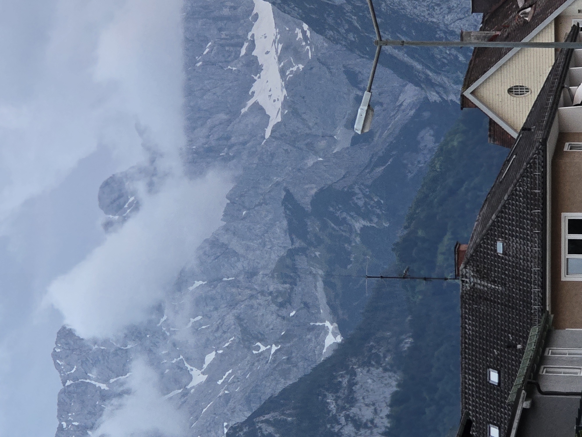 The image captures a view of the Zugspitze massif, Germany's highest peak, from the town of Garmisch-Partenkirchen. The mountains, partially obscured by dramatic clouds and streaks of snow, dominate the background. In the foreground, traditional Bavarian-style houses with tiled roofs are visible, interspersed with a street lamp and an antenna. The overall atmosphere is overcast and moody, suggesting a cool, perhaps rainy, day. There are no people or animals depicted in the image.