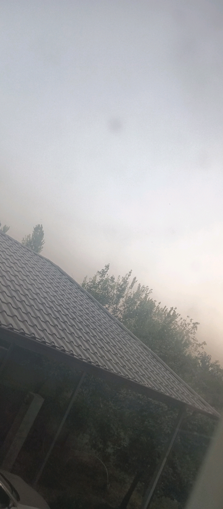 This image shows a hazy sky and a portion of a dark, tiled roof in the foreground. Trees with green foliage are visible in the background, partially obscured by haze. The overall scene suggests a possibly smoky or foggy atmosphere. No people or specific actions are discernible. The location is identified as Somoniyon, Tajikistan.