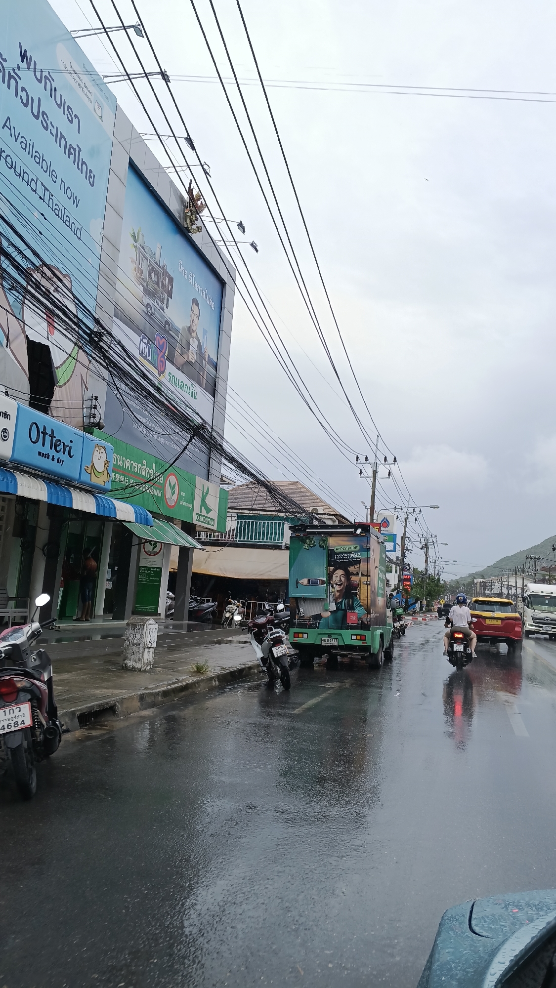 The image captures a street scene on what appears to be a wet day in the Ko Samui District of Thailand. The foreground shows a wet asphalt road with reflections of vehicles and overhead lights. On the left side of the road, several motorcycles are parked along the sidewalk. A person is visible under the awning of a shop labeled "Otteri wash & dry" and another further down the street.
The middle ground features a street with several vehicles. A green truck with advertisements on its side is parked, and a person is seen standing near it. Two motorcycles are on the road, one parked and one moving. A red car and a white truck are also visible in traffic. Overhead, a complex network of power lines stretches across the frame.
The background includes buildings lining the street, with signage that suggests a commercial area. A large billboard with Thai text and images is prominently displayed on the left. Beyond the buildings, a lush green hillside is visible under a cloudy sky. The overall impression is of a bustling urban environment in a tropical location, possibly experiencing or recovering from rain.