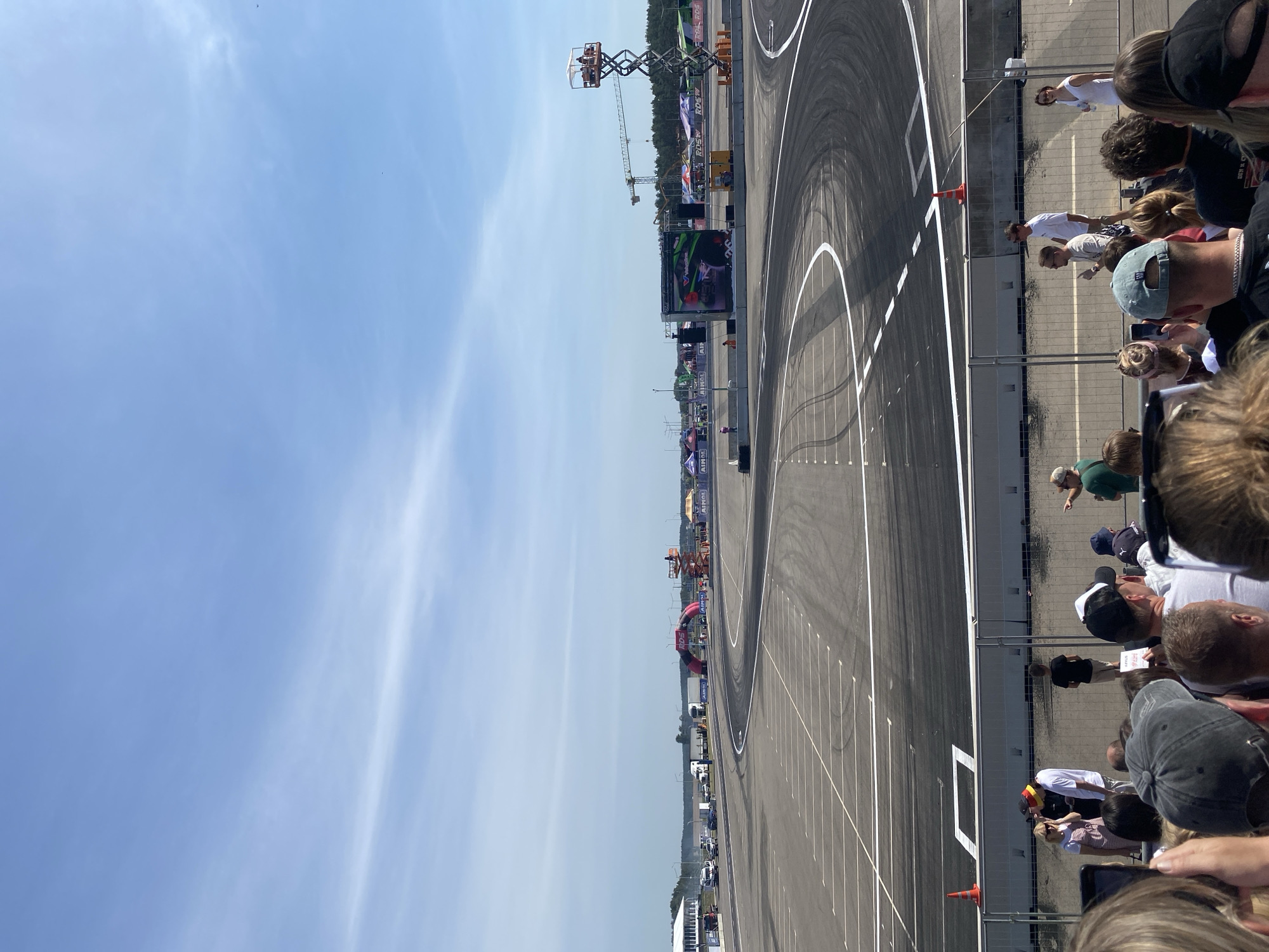 The image shows a wide, paved race track with white lines marking lanes and curves. In the background, there are several tents and structures, some with event banners. A large inflatable archway, red and black, is visible in the distance, with "RDS" lettering on it. To the right, a scissor lift is positioned with people on top, and a construction crane is also present. A large screen displays a racing image. In the foreground, a crowd of spectators stands behind a metal fence, watching the event. Several individuals are wearing hats, and some are holding up phones. The sky is blue with scattered clouds. The location appears to be a race track or a similar outdoor event venue. Based on the "RDS" branding, this is likely a drift racing event. While the image does not explicitly state the location as Minsk, Belarus, RDS (Russian Drift Series) events have been held in various locations across Eastern Europe, including Belarus.