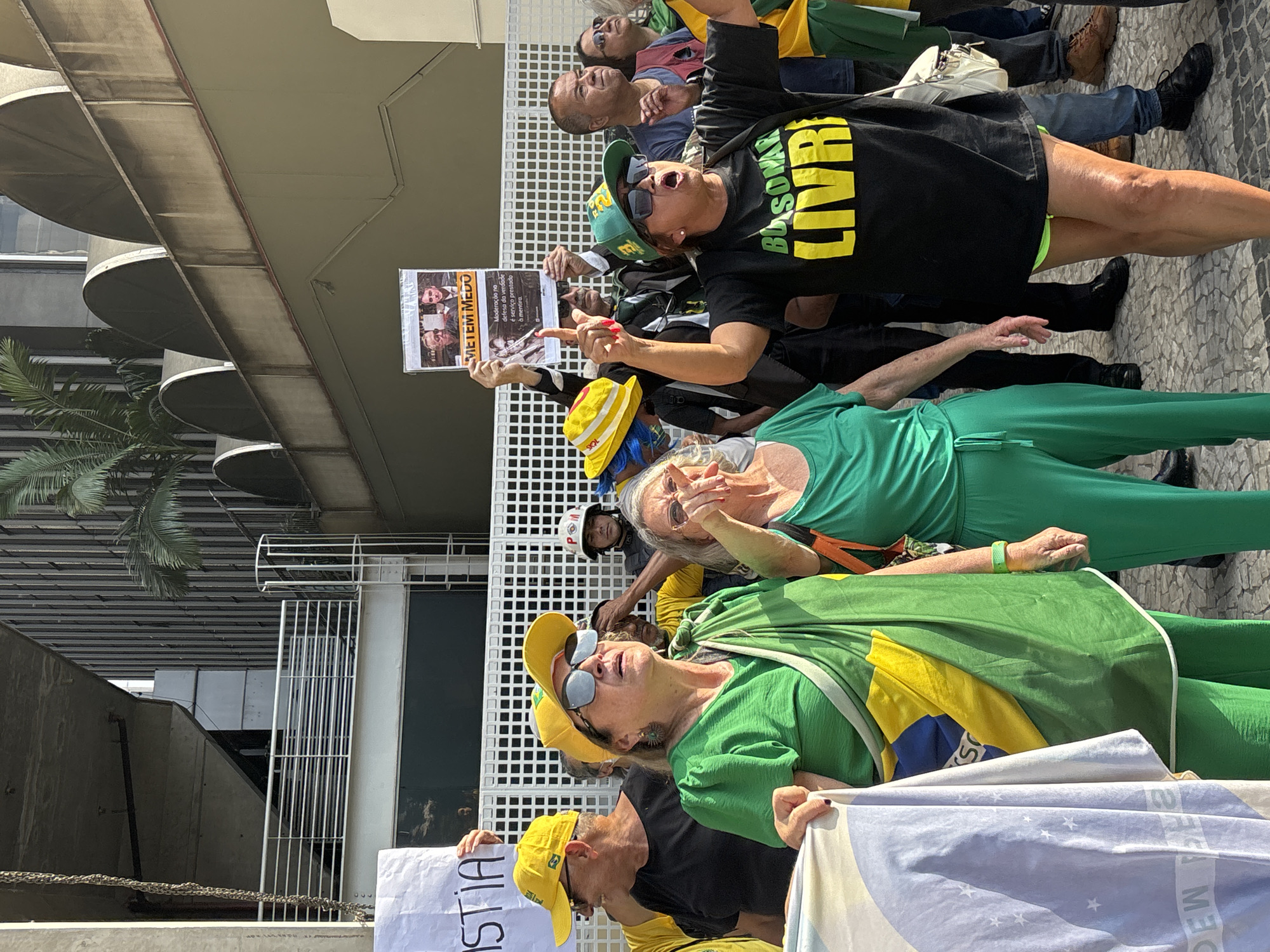 A group of people, predominantly women, are gathered outdoors under bright daylight, likely participating in a political protest or rally in São Paulo, Brazil. The scene is set against a backdrop of modern building facades with curved balconies and a grid-like fence.
The individuals are expressive and animated, with some raising their arms, pointing, and looking up. Many are wearing clothing in shades of green and yellow, the colors of the Brazilian flag, which are often associated with conservative political movements in the country. Several people are wearing green and yellow caps, and one individual is draped in a Brazilian flag.
Notable clothing includes a black t-shirt with the yellow text "BOLSOMAR LIVRE" (Bolsonaro Free), and a person wearing a full green jumpsuit. Sunglasses are worn by several attendees.
Visible text on signs and banners indicates the nature of the protest. A white banner at the bottom left shows "ISTIA...", likely part of the word "JUSTIÇA" (Justice). A prominent sign held aloft features images of two men, one identifiable as Jair Bolsonaro, with the bold text "METEM MEDO" (They Scare/Intimidate). Below this, smaller text reads: "Moderação na defesa da verdade é serviço prestado à mentira" (Moderation in defense of truth is a service rendered to lies). The text "DEW" and "S" are also partially visible on a Brazilian flag.
The overall atmosphere suggests an impassioned public gathering, with participants actively demonstrating their political views.