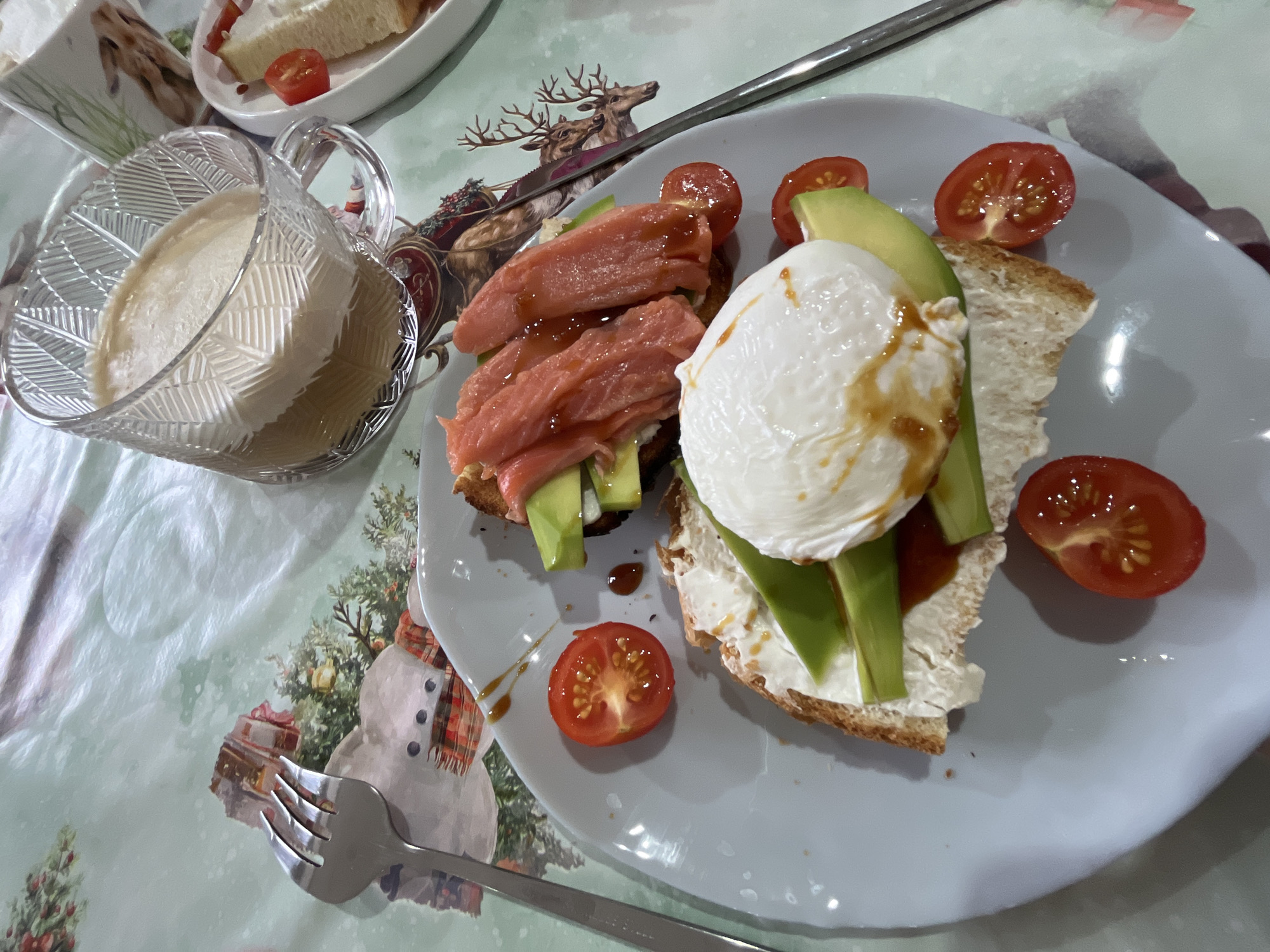 The image shows a meal consisting of two open-faced sandwiches on a light gray plate. One sandwich has a creamy spread, avocado slices, and a poached egg drizzled with a dark sauce. The other has avocado slices and salmon drizzled with a dark sauce. Cherry tomato halves are scattered around the sandwiches. To the left of the plate is a glass mug filled with a light-colored liquid and a solid, white mass. A fork lies next to the plate. A second plate is visible in the upper left corner, containing what appears to be bread and a cherry tomato. The tablecloth features a winter theme with images of reindeer, a Christmas tree, and a snowman. The location is Chaikovsky, Russia.