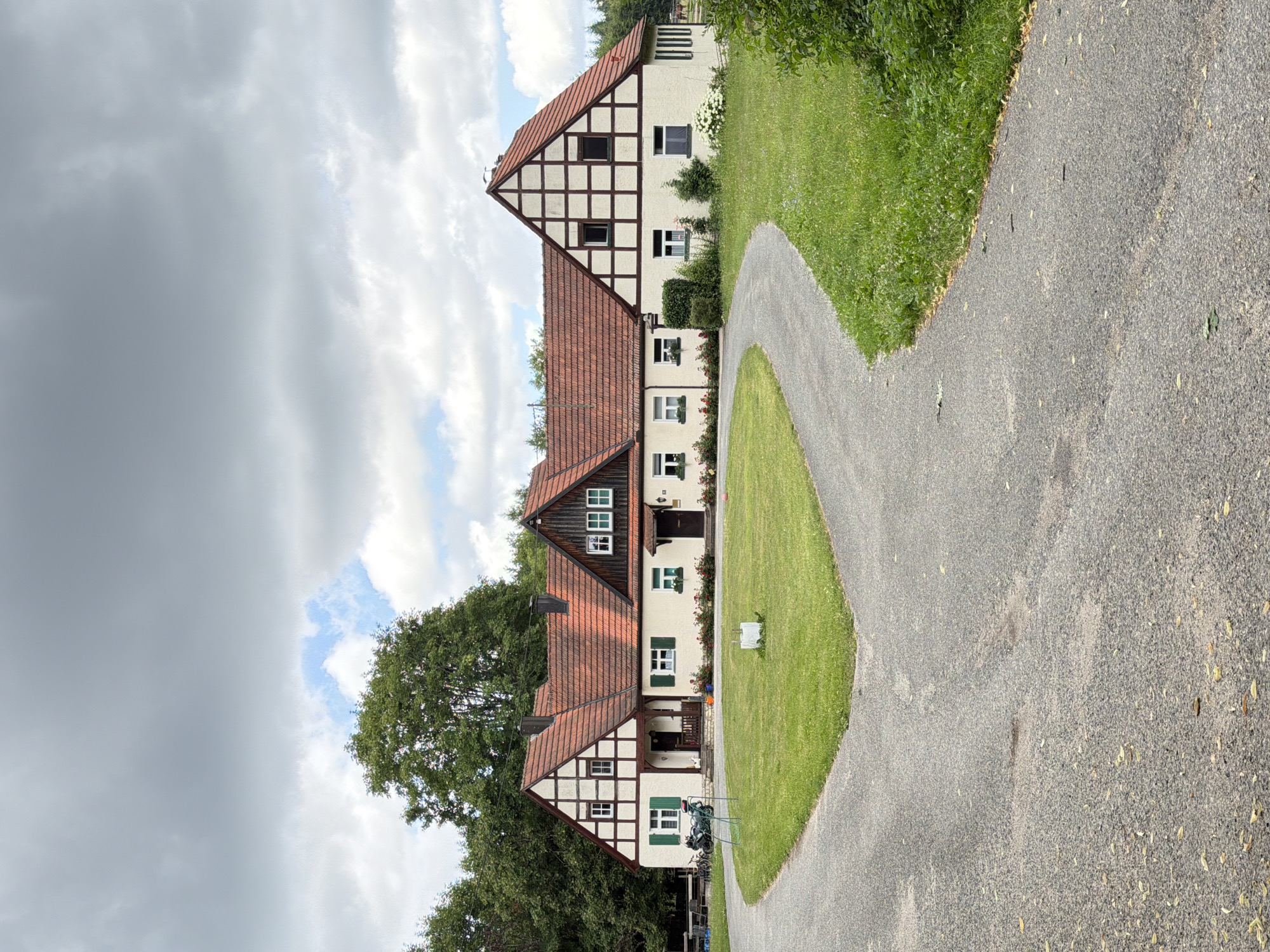 The image shows a large, two-story building with a traditional German half-timbered architectural style, featuring a red tile roof and white stucco walls with dark wooden beams. The building is set in a rural environment, with a large, well-maintained lawn in front. A gravel driveway splits and curves around the lawn, leading to the building. Trees and greenery surround the property, with a dense forest visible in the background. The sky is overcast with dramatic clouds. There are no people visible in the image. The building appears to be a residence or possibly a farmstead. The location is identified as Heiligenstadt i. OFr., Germany.