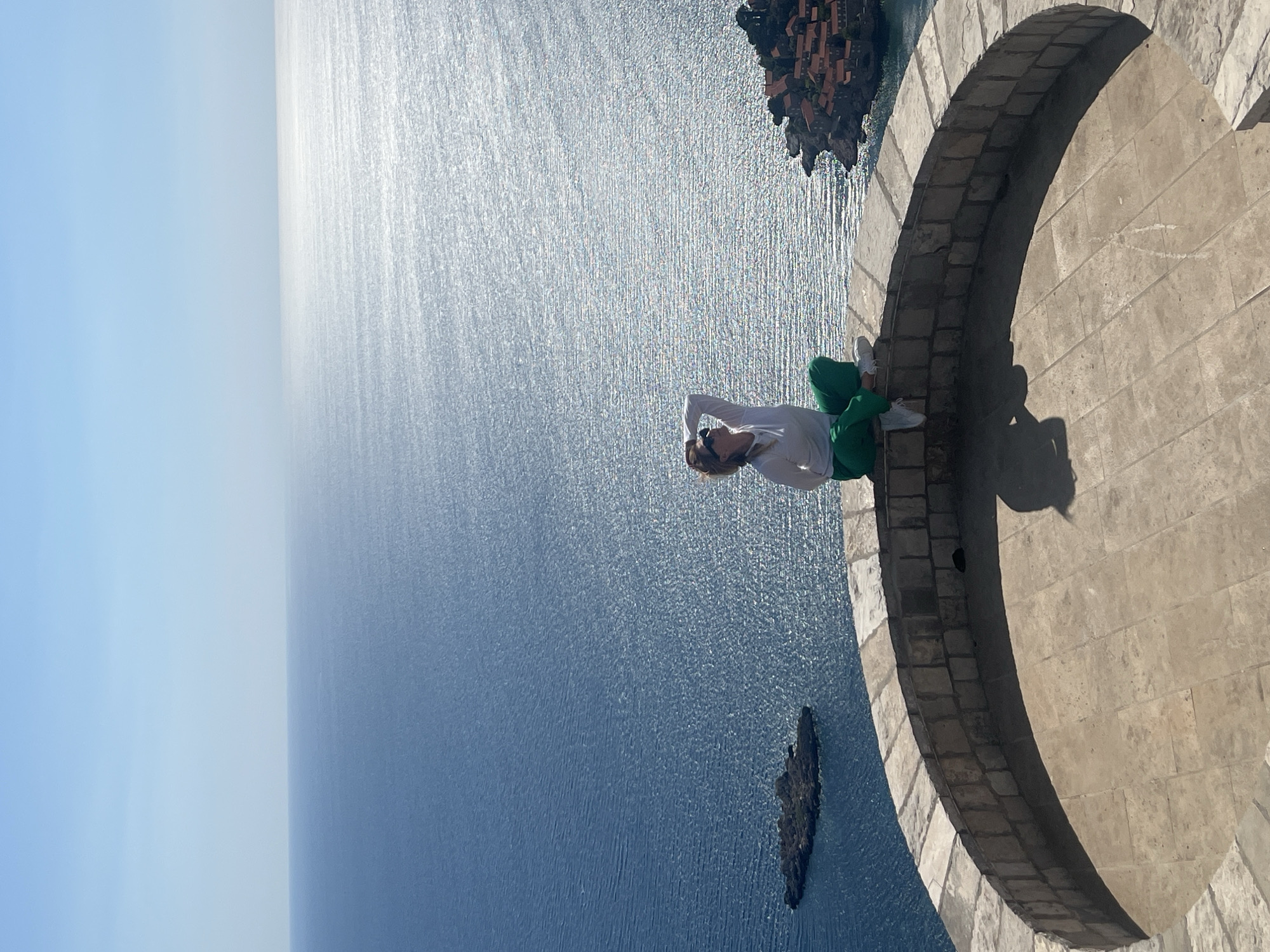 The image presents a breathtaking coastal view from a high vantage point, dominated by a vast expanse of sparkling sea under a bright sky. In the foreground, a person, identified as a woman, stands on a curved, rustic stone balcony or wall. She is positioned with her back partially to the viewer, looking out towards the horizon. She wears a white long-sleeved top, green pants, and white sneakers, with sunglasses on and her hands resting behind her head, indicating a relaxed posture as she takes in the expansive scenery.
The sea, a brilliant blue, stretches far into the distance, with strong sunlight creating a dazzling, shimmering effect across its surface. This reflection makes parts of the water appear intensely bright, contrasting with deeper blue tones elsewhere. In the mid-ground, a prominent rocky island or promontory, densely covered with terracotta-roofed buildings, is visible, anchoring the scene. This distinctive landmark identifies the location as Sveti Stefan in Montenegro. A smaller, darker rock formation also dots the water closer to the viewpoint.
The time of day is clearly sunny daytime, likely midday or early afternoon, given the intensity of the sun's reflection on the water and the deep shadows cast by the stone structure and the person onto the wall. The weather is clear and bright, contributing to the serene and awe-inspiring atmosphere of the scene. No visible text is present in the media.