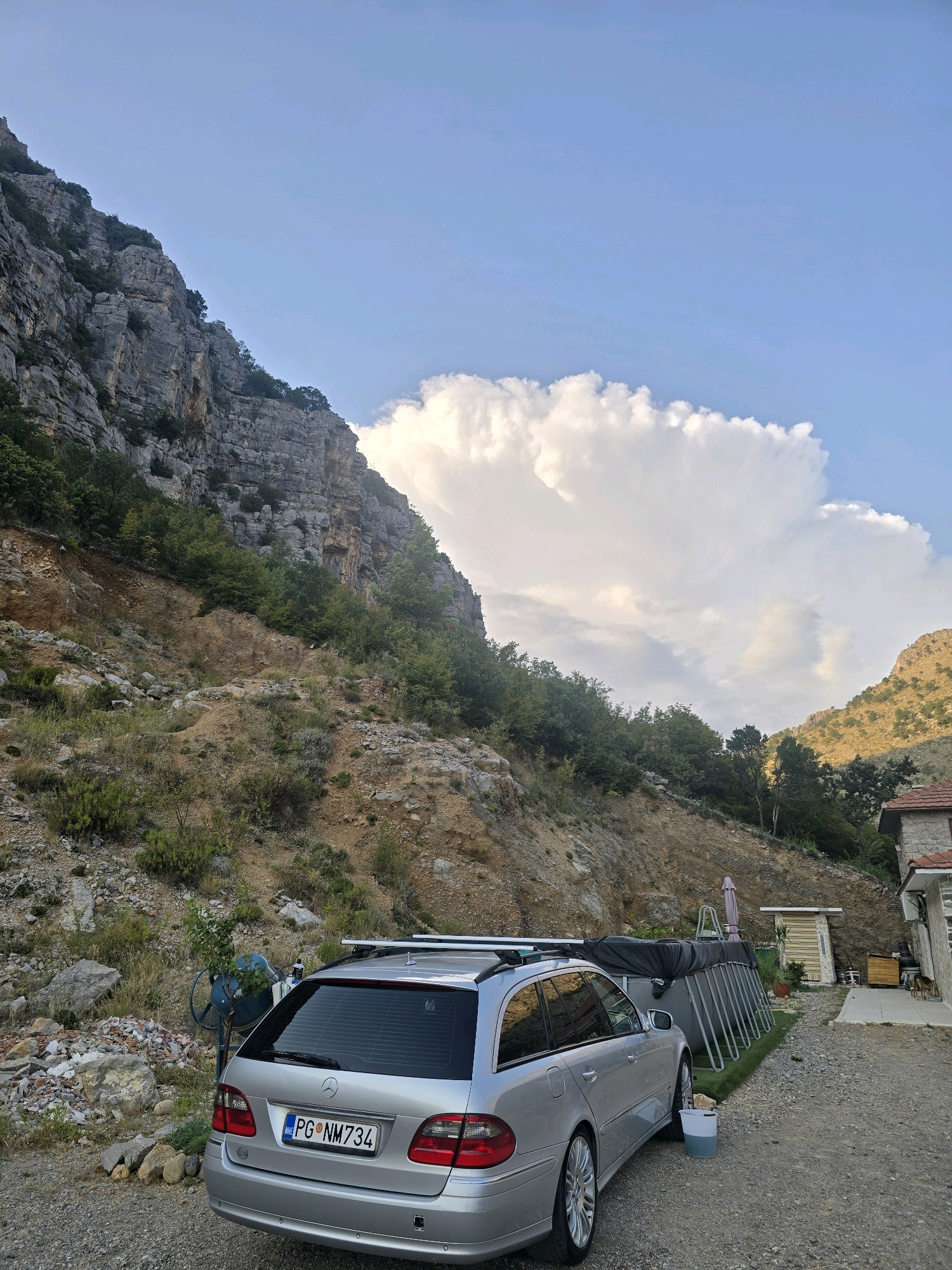 This image captures a scene in Podgorica, Montenegro, characterized by a dramatic mountainous backdrop and a residential area. A silver Mercedes-Benz E-Class station wagon is parked on a gravel path in the foreground. To the right of the car, an above-ground swimming pool is set up, partially covered. Adjacent to the pool is a small building with a light-colored facade and a tiled roof. Further to the right, a stone house with a red-tiled roof is visible, with a dog standing near its entrance. The left side of the image is dominated by a steep, rocky mountain slope covered with sparse vegetation and trees. A large, white cumulus cloud formation is prominent in the sky above the mountains. The overall impression is one of a rural or semi-rural setting within the mountainous terrain surrounding Podgorica.