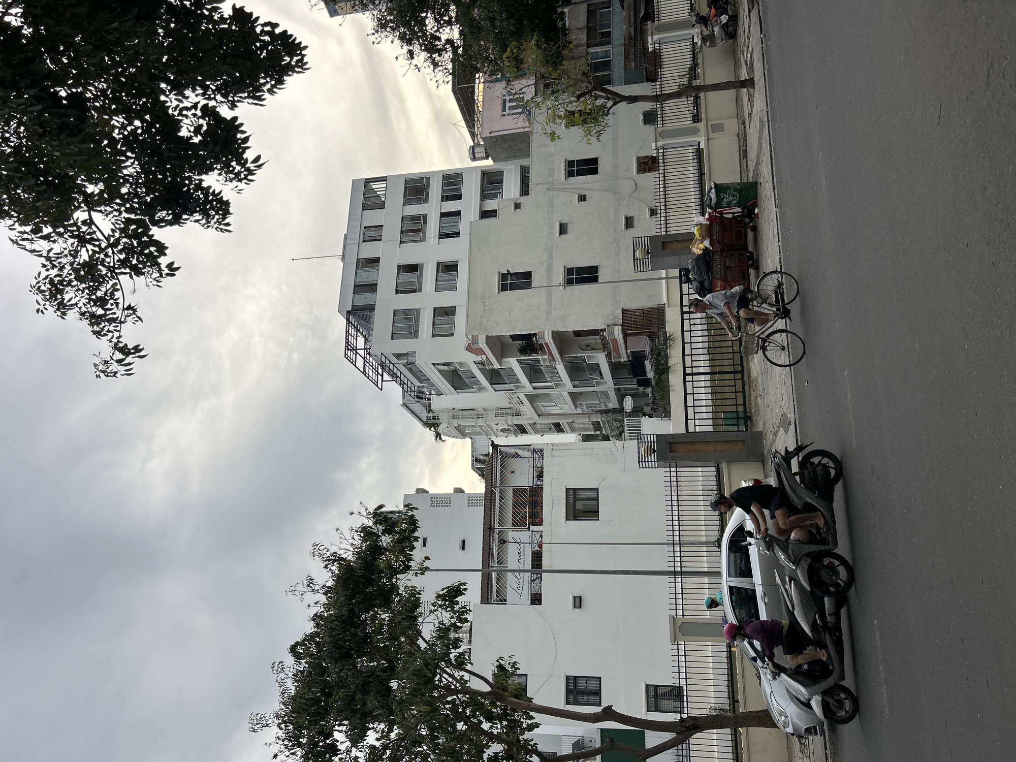 The image depicts an urban street scene in Nha Trang Ward, Vietnam, on an overcast day. The perspective looks down a street lined with multi-story buildings and trees.
On the left side of the frame, a row of predominantly white, modern-style buildings features numerous windows and balconies, some with dark metal railings or grilles. A large tree with dense, dark green foliage partially obscures the top left and right portions of the buildings. The sky above is cloudy and light grey.
The paved street runs from the foreground towards the background, parallel to the buildings. A man wearing a striped shirt, shorts, and a cap is cycling a bicycle away from the viewer on the road. Further along the curb, a light-colored sedan is parked, with two motorbikes positioned next to it. One person, dressed in a dark shirt and shorts, is seen with the front motorbike, while another person wearing a purple top and a pink helmet sits on the second motorbike.
Along the sidewalk, black metal fences border the properties, and two green trash bins are visible near one of the fences. One building has a sign with the text "Lelenady Beauty" visible on it. The scene portrays typical daily life on a Vietnamese street.