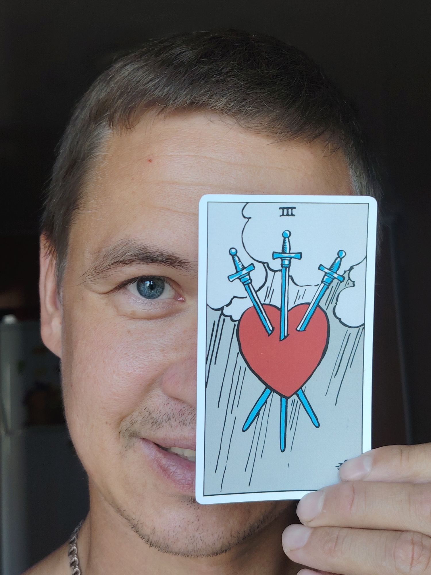 A man is holding a tarot card, the Three of Swords, in front of the right side of his face, obscuring his eye and nose. He has light skin, short brown hair, and blue eyes. The tarot card features a red heart pierced by three blue swords, with clouds and rain depicted in the background. The man is wearing a silver chain necklace. The background is dark and out of focus, with hints of a refrigerator and possibly other kitchen appliances visible on the left side. The scene is indoors, likely in a residential setting in Chaikovsky, Russia.