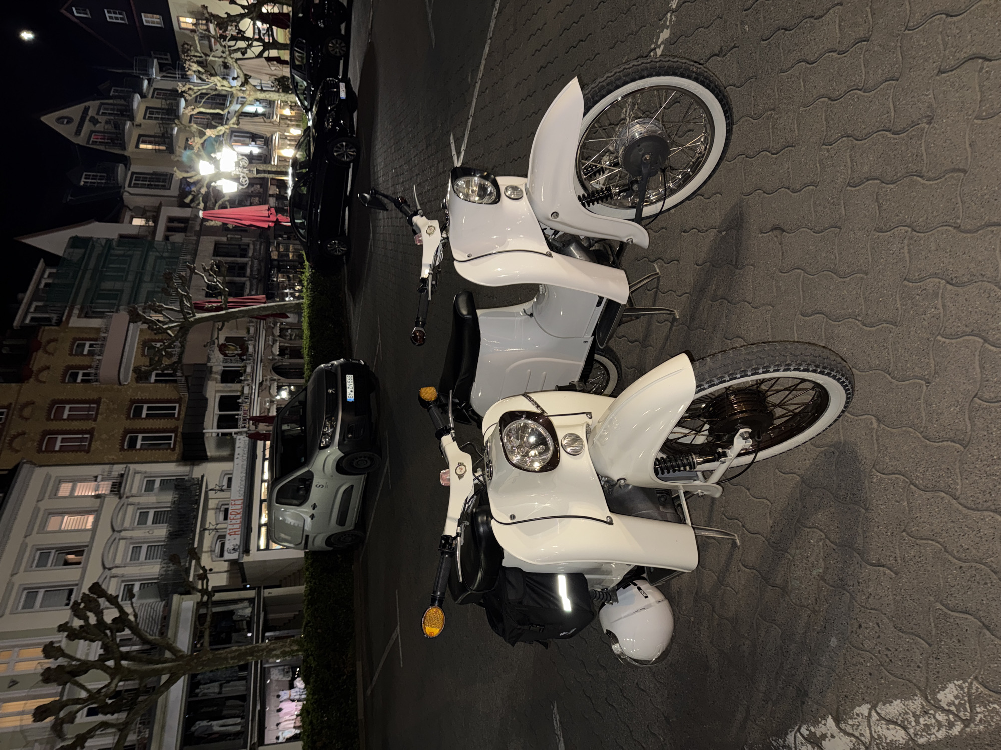Two white scooters are parked on a paved surface in Cochem, Germany. The scooters are facing opposite directions. Buildings with lit windows are visible in the background, along with parked cars. The setting is at night.