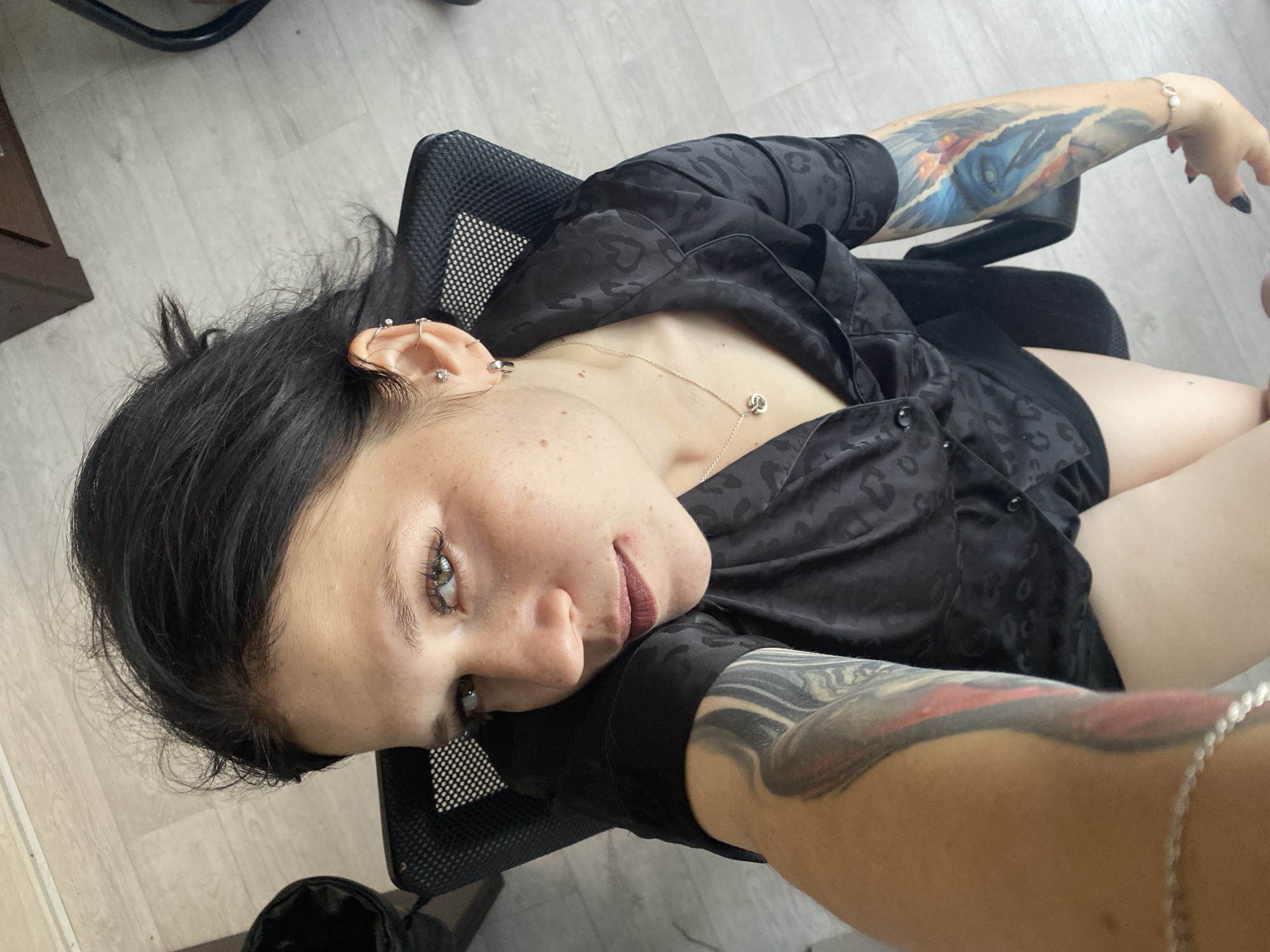 A young woman with dark hair, green eyes, and freckles is seated in a black office chair. She is wearing a black, patterned silk shirt and has tattoos on both arms. One tattoo on her left arm is a black and grey abstract design, while the tattoo on her right arm is a colorful depiction of a face, possibly a character from a movie or game, with blue and red elements. She is looking directly at the camera with a slightly open mouth. The background appears to be an indoor setting with light-colored wooden flooring and a dark piece of furniture in the upper right corner. There is a black trash can visible on the left side of the frame. There is no information in the image that would indicate the location within the city of Chaikovsky, Russia.