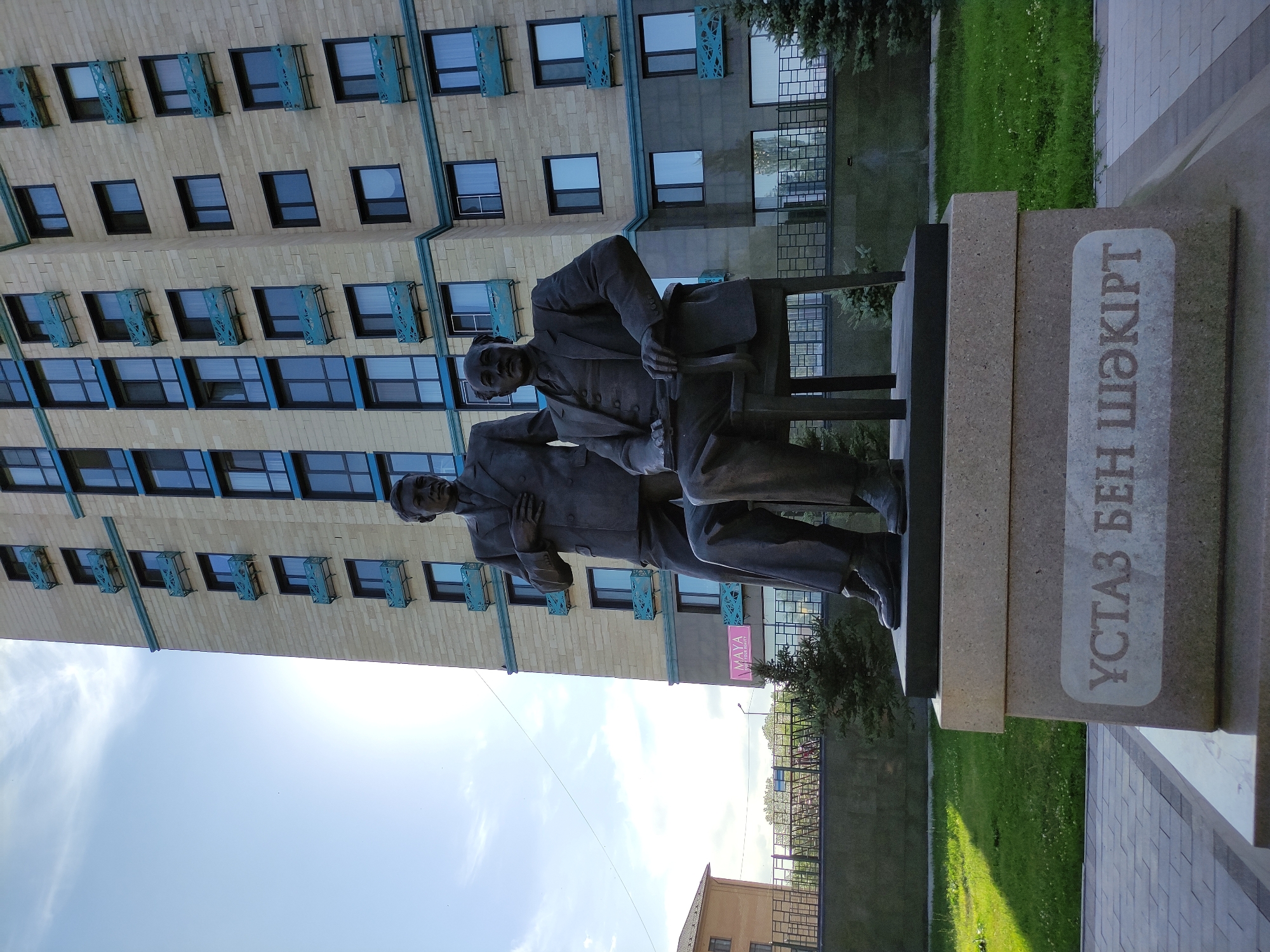 The image shows a bronze statue of two men in suits. One man is standing and has his hand on his chest. The other man is seated in a chair, holding a book or tablet. Both men are looking in the same direction. The statue is placed on a stone pedestal with Cyrillic writing that reads "УСТАЗ БЕН ШӘКІРТ". In the background, there is a tall, modern apartment building with beige brickwork and blue window frames. There is also a patch of green grass and some trees. The photograph was taken on a sunny day with a clear sky. The statue is located in Semey, Kazakhstan, and it is a monument to the teachers of Abai.