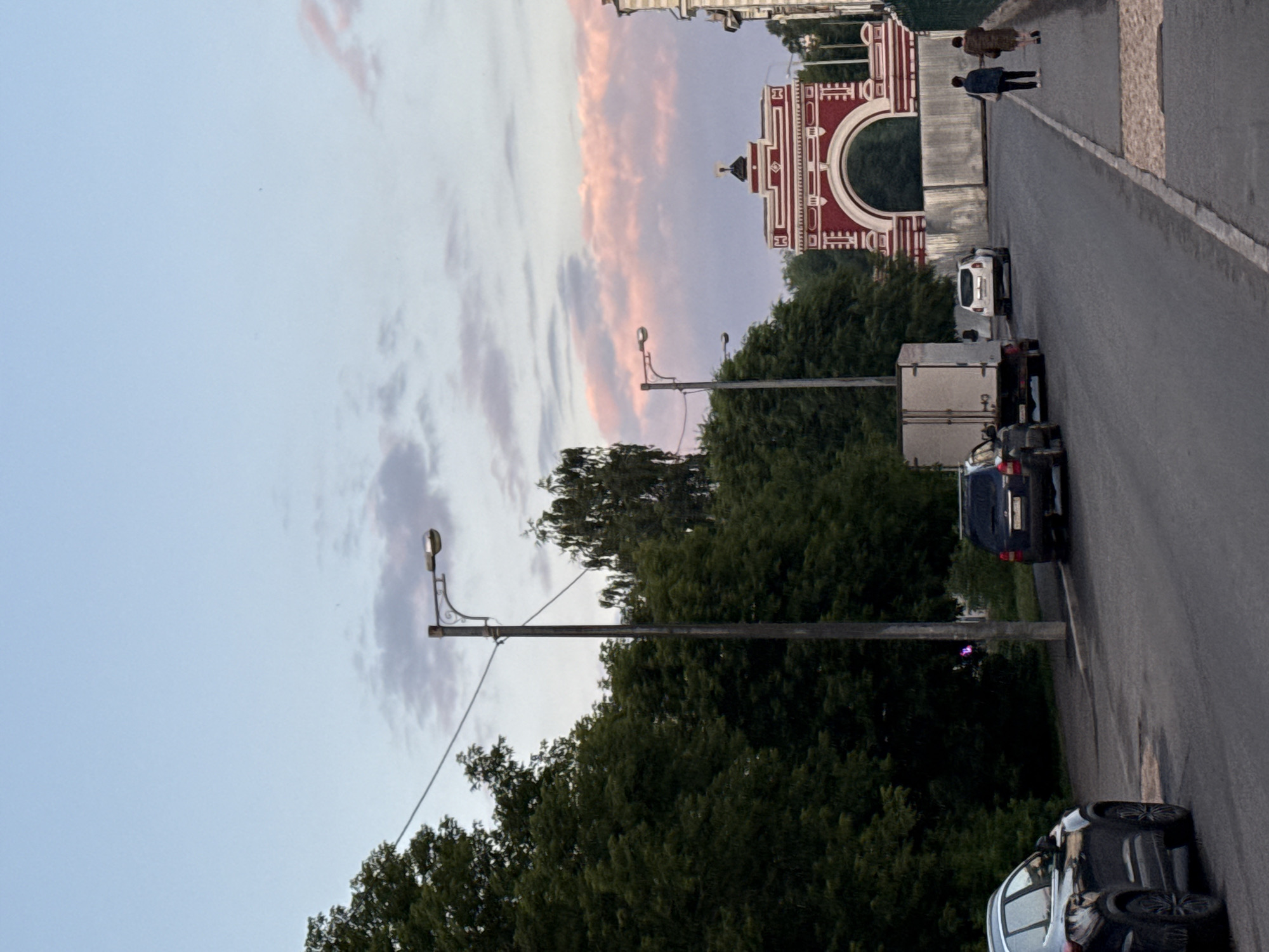 A street scene unfolds in Kazan, Russia, under a twilight sky painted with soft pink and blue hues. The perspective is from the side of a paved road, with a sidewalk on the right. Dense green trees line the left side of the road, creating a shaded canopy. A dark-colored SUV is parked on the left side of the road, facing away from the viewer. Further down the road, a white box truck is parked behind a dark-colored car.
Dominating the background is a strikingly red and white arched structure, resembling a gateway or a monumental arch, with intricate decorative elements. Two people, a woman in a dark jacket and a woman in a patterned coat, walk along the sidewalk on the right, moving away from the viewer. The overall atmosphere is serene, capturing a moment in the early evening in a Russian city.