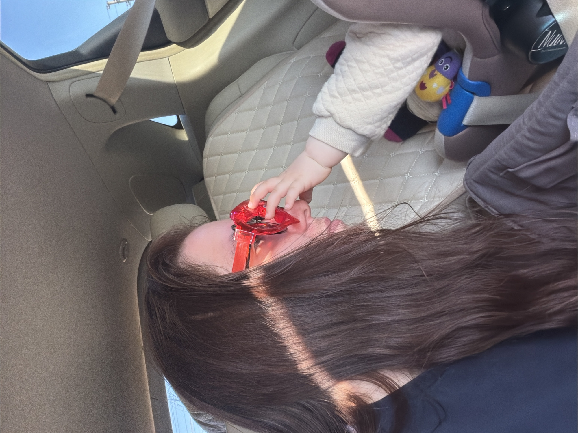 The image shows a woman with long brown hair wearing red sunglasses, possibly inside a car. A baby's hand reaches towards her face. The baby is seated in a car seat, and a soft toy is attached to the car seat. The car interior has light-colored upholstery. The location is not apparent from the image.