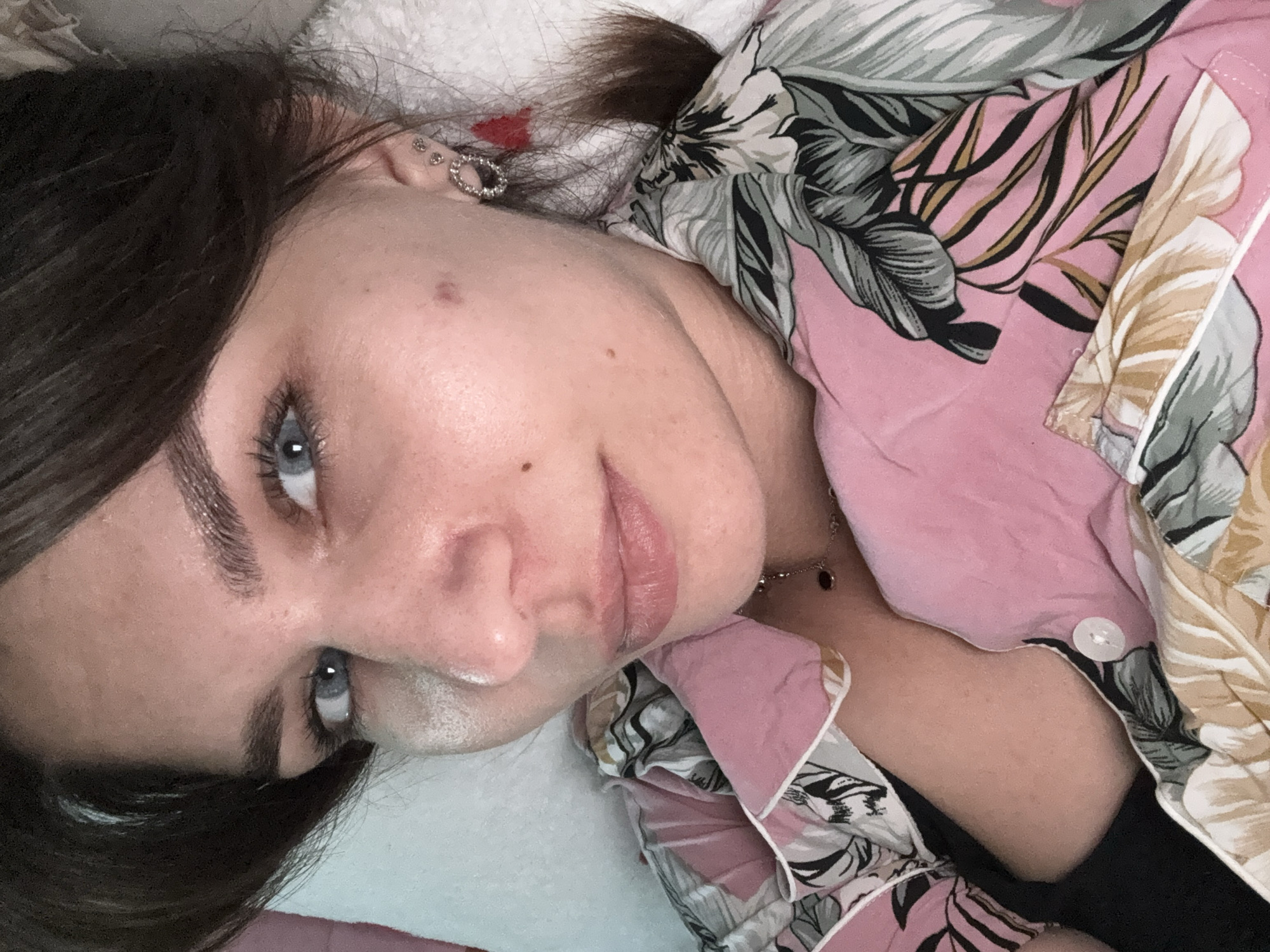 A close-up, high-angle selfie shows a woman with brown hair and blue eyes lying down. She is wearing a pink pajama top with a tropical leaf pattern. Her head is tilted slightly to her left, and she is looking at the camera. She has a small mole on her left cheek and is wearing hoop earrings. A dark necklace is visible around her neck. The background appears to be a white pillow and blanket.
