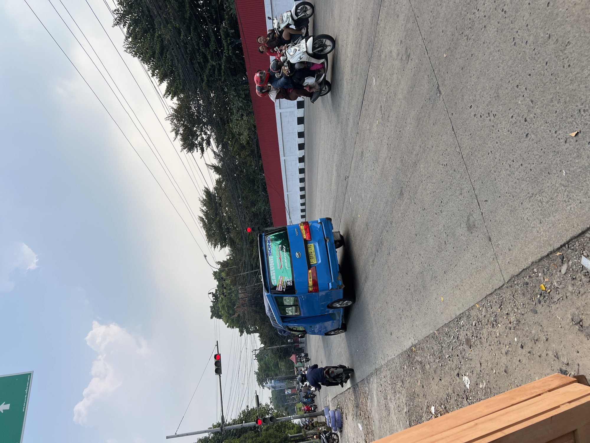 The image depicts a street scene in Citeureup, Indonesia. A blue van with "GARUNI SONG" written on the back travels down the right lane of a paved road. Multiple motorcycles are also present on the road, with two on the right side carrying passengers, and several more on the left side, some parked. A traffic light on the left side of the road is red. Trees and utility lines line the sides of the road, with a red and white striped wall in the background on the right. The sky is overcast with some scattered clouds. The ground consists of asphalt and gravel. A wooden structure is visible in the foreground on the left.