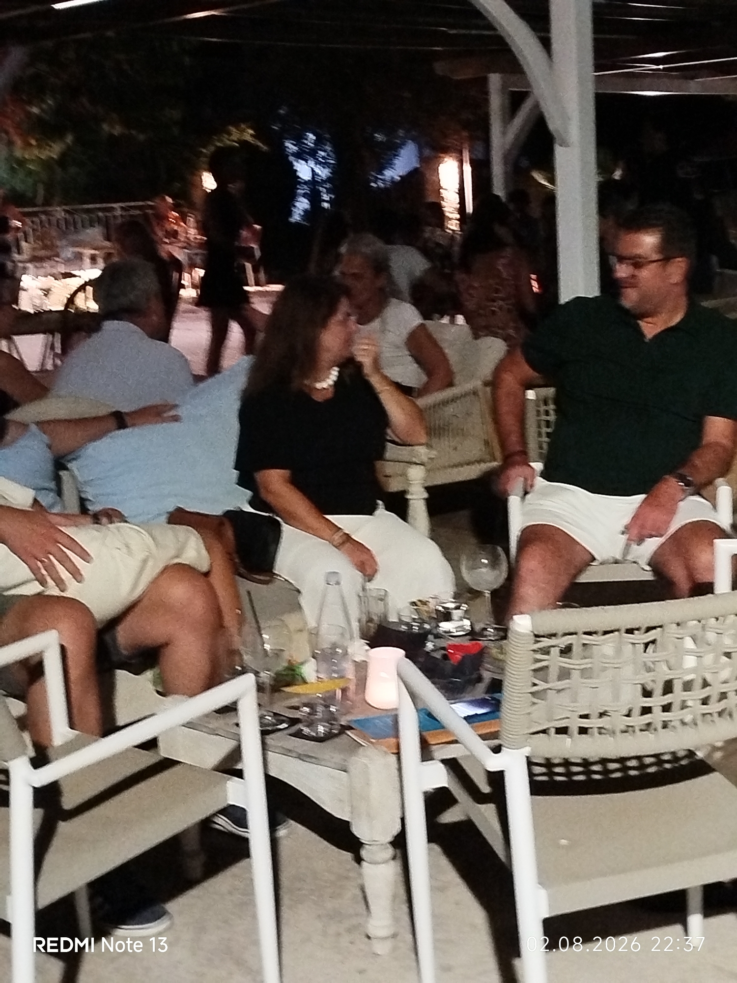 A medium shot captures a group of people seated at tables outdoors, likely at a restaurant or cafe in Sithonia Municipal Unit, Greece. The scene appears to be illuminated by ambient lighting, creating a dimly lit atmosphere. In the foreground, a man wearing a dark green polo shirt and white shorts is seated, looking to his left. To his right, a woman in a black top and white pants is visible, her head turned downwards. In the mid-ground, another man in a light blue shirt is seated, with his arm resting on the table. A person in a light-colored shirt and shorts is also seated in the foreground, with their legs crossed. Various objects such as glasses, a bottle, and a small table are present. The background shows trees and other patrons, suggesting a lively setting. The image is dated 02.08.2026 and has a time stamp of 22:37.