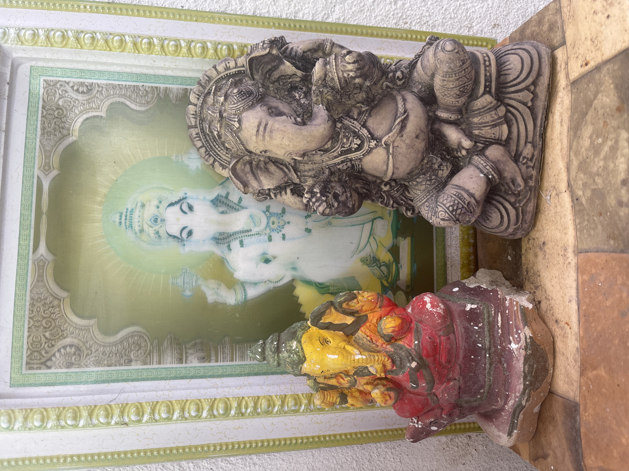 The image depicts a vertical arrangement of objects against a wall in Thalaramba, Sri Lanka. At the back is a framed picture with a light green and yellow border. The picture features a depiction of the deity Ganesha in soft, pastel colors.
In front of the picture are two Ganesha statues. The upper statue is a grey, stone-like figure of Ganesha lying down. Below, a smaller, more colorful statue of Ganesha is present, painted with shades of yellow and red. The statues appear to be positioned outdoors. The lower portion of the scene includes tiled or stone surfaces. The wall itself is rough and slightly off-white.