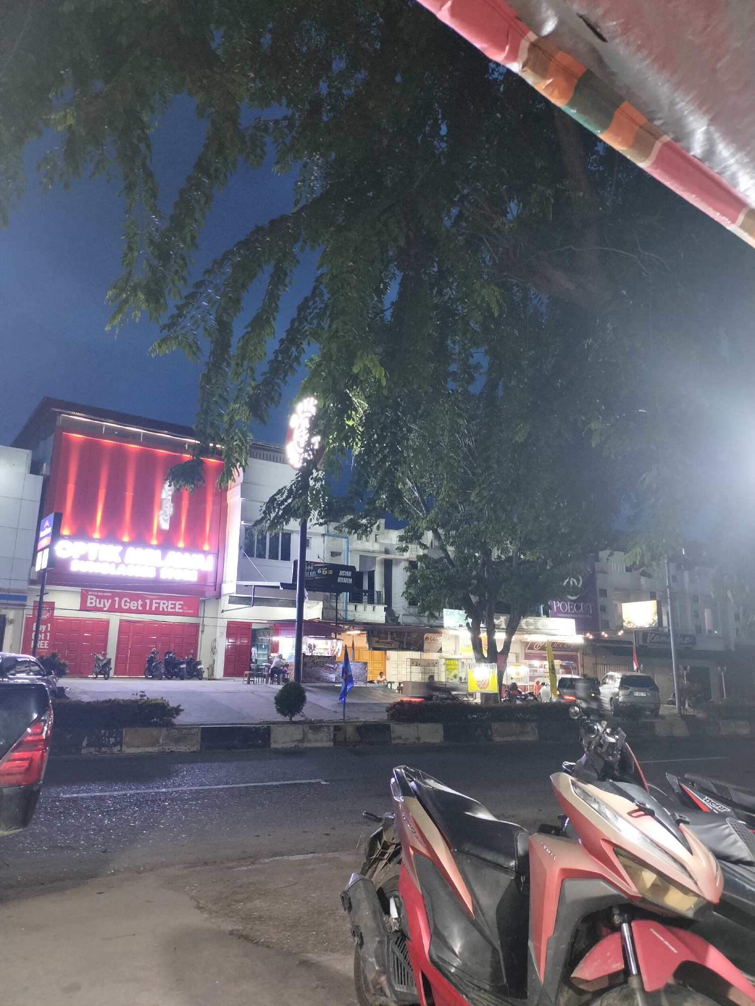This is a nighttime street scene in Langsa, Indonesia, likely captured from across the street, looking towards a row of businesses. A large tree with dense foliage partially obscures the view of the buildings. The most prominent structure is a building with a bright red facade, advertising "OPTİK MIL ALAM SUNGLASSES STORE" with illuminated text. Below the store name, a banner announces "Buy 1 Get 1 FREE." Multiple red garage doors are visible on the ground floor of this building.
To the right of the red-fronted building, other businesses are visible, some with illuminated signs. One sign reads "POECUT." A street lamp with bright illumination is visible on the right side of the image, casting light across the scene. In the foreground, the street surface is wet and reflective, suggesting recent rain. Several motorcycles are parked along the sidewalk in front of the businesses, and two motorcycles are in the immediate foreground, with one a prominent red and gold color. A few individuals are seated at tables outside one of the establishments. The sky is dark, indicating nighttime. The overall impression is a lively commercial street at night.