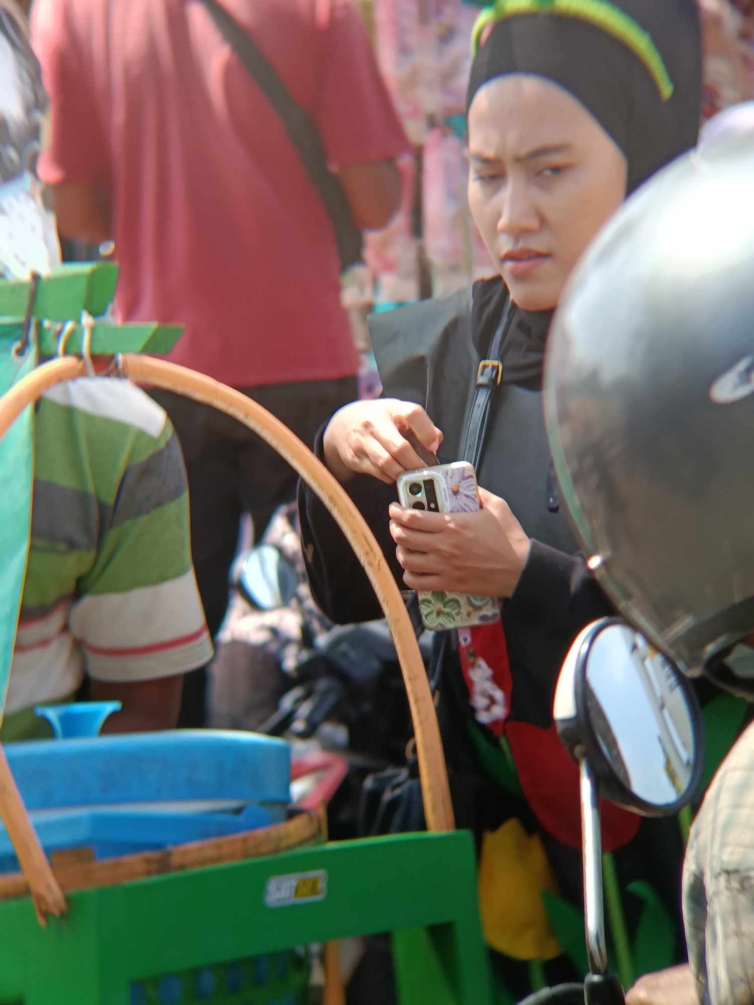 In Desa Kebon Manis, Kecamatan Cilacap Utara, Indonesia, a person wearing a black hijab with a yellow and green band is holding a mobile phone with a floral case. The person is wearing a black outer garment and has a black strap across their body. In the background, another person is wearing a red t-shirt. A green cart with a blue container is visible in the foreground, with a person wearing a striped shirt partially visible behind it. A motorcycle helmet is also present in the image, with its mirror reflecting the surroundings. The scene appears to be outdoors, likely on a street or market.