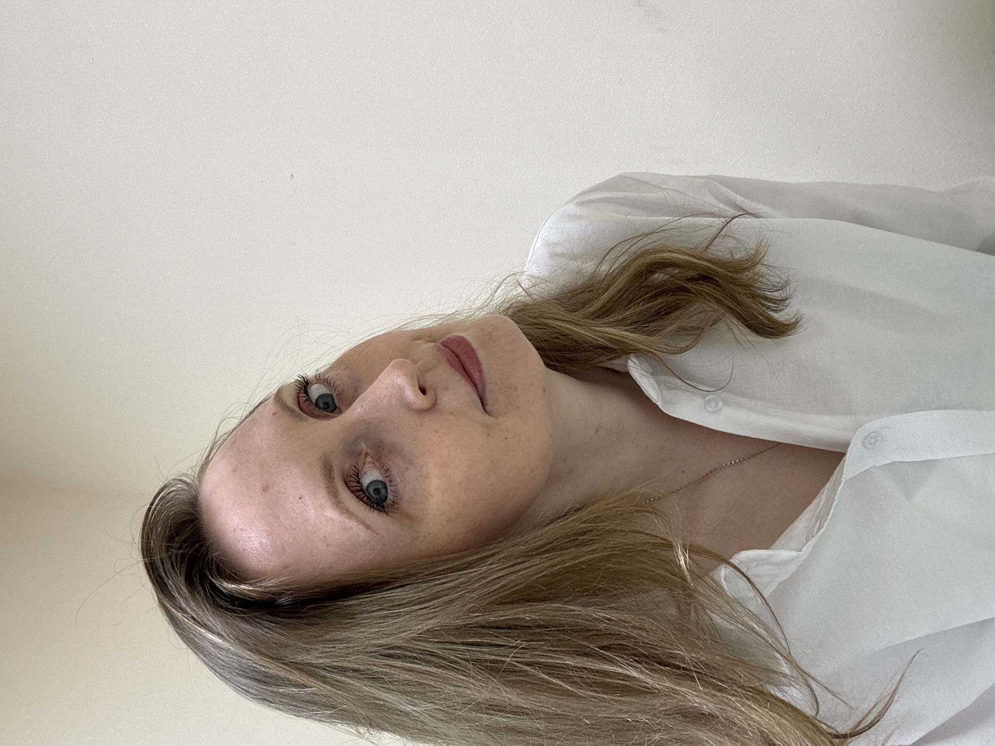 A Caucasian woman with blonde hair and blue eyes is pictured from a slightly low angle, rotated counter-clockwise. She is wearing a white collared shirt that is partially buttoned. A gold chain necklace is visible. The background is a plain, light-colored wall. No elements indicate the location is Chaikovsky, Russia.