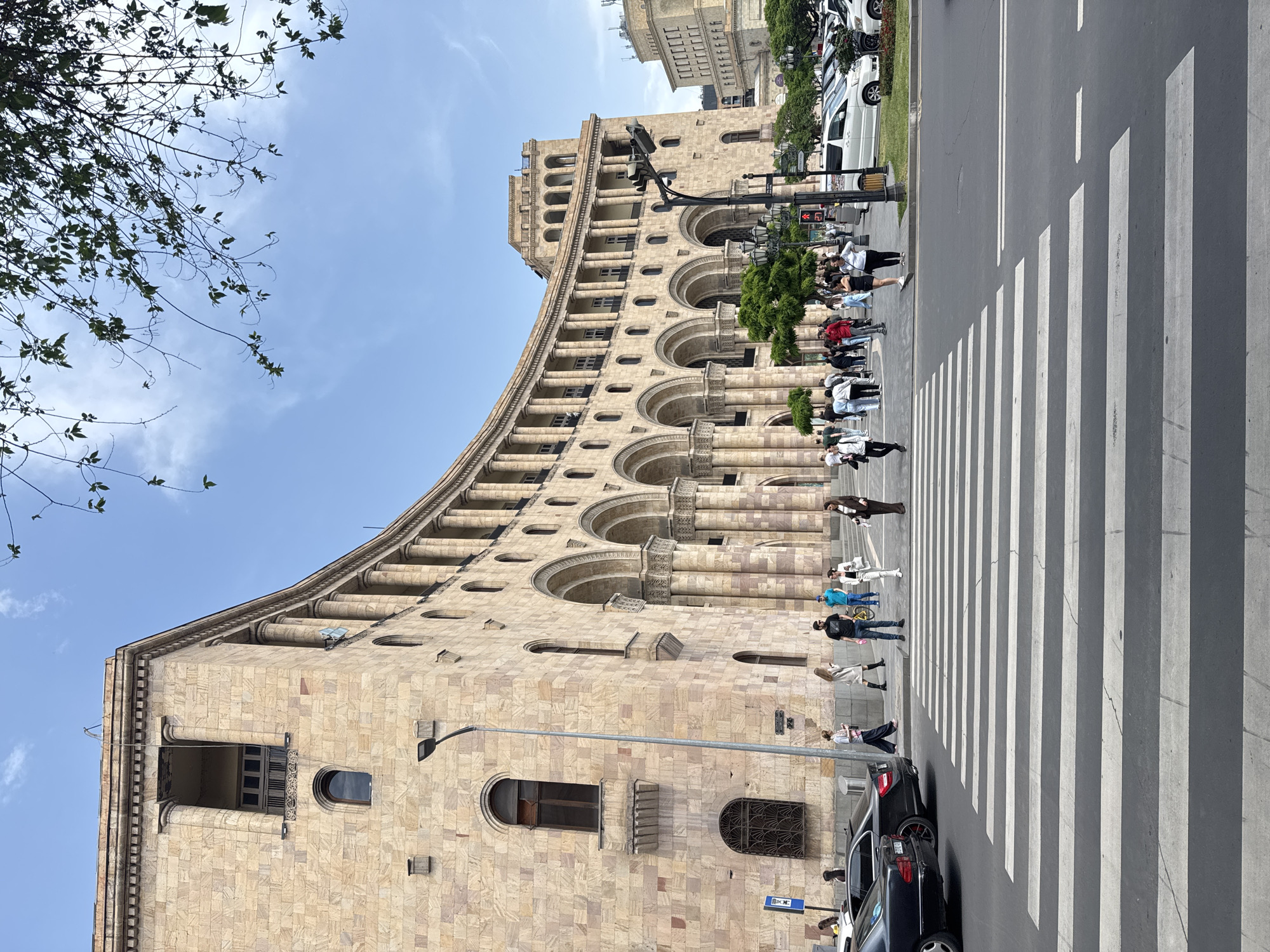The photograph captures a street scene in Yerevan, Armenia, featuring a large building with a light-colored stone façade and a series of arched windows and columns. A pedestrian crosswalk is in the foreground, with several individuals in casual attire crossing the street. Some are walking alone, while others are in small groups. Cars are parked along the curb on the right side of the frame. A streetlight is visible in front of the building, and some tree branches are visible in the top left corner of the image. The sky is light blue with a few clouds. The architectural style of the building is likely Soviet Modernism. The location appears to be Republic Square, Yerevan, Armenia.