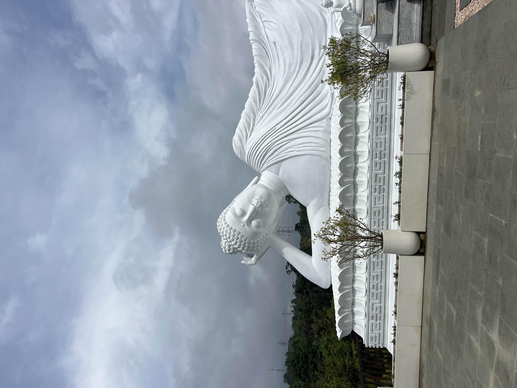 A white, reclining Buddha statue rests on its left side, with its head supported by its left hand. The statue is positioned outdoors against a backdrop of a cloudy sky and a dense green forest. Power lines are visible in the background. The statue rests on a platform adorned with ornate geometric designs, and planters with small trees line the base. Gray stone tiles cover the ground around the statue. The photo was taken in Pupuan, Indonesia.