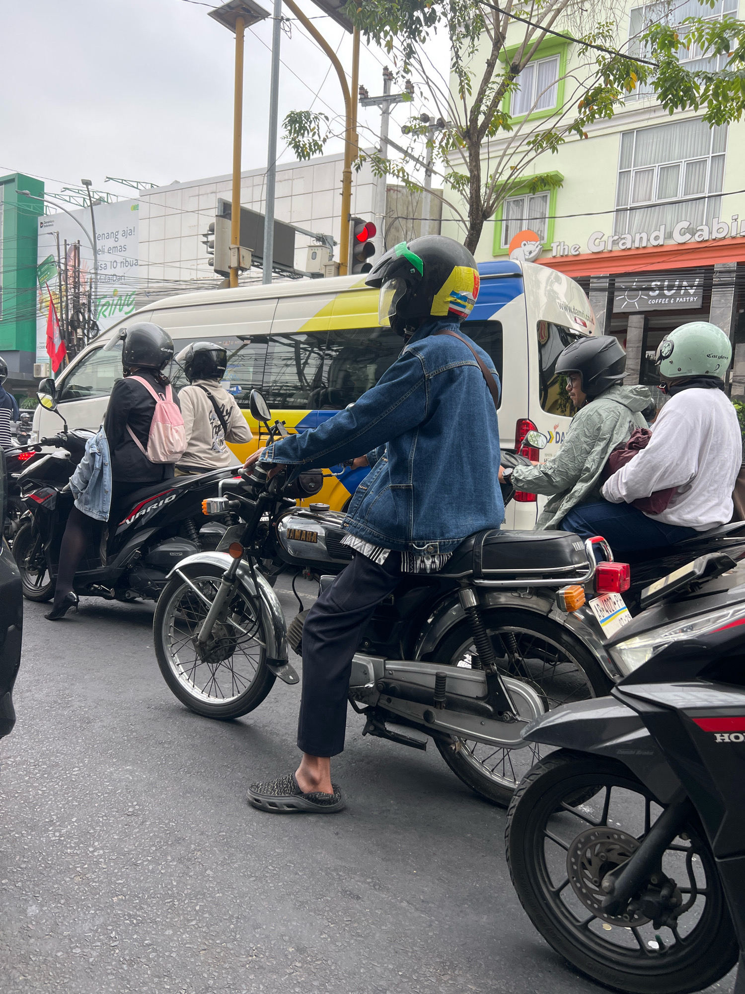 The image captures a street scene in Yogyakarta, Indonesia, likely during daylight hours. Several motorcycles, some with riders, are present on a paved road. The rider in the foreground is wearing a denim jacket and black pants, with a black helmet. A second motorcycle to the left has two riders; the one in front wears a black jacket and a pink backpack, while the one behind has a white hoodie and a black helmet. To the right, another motorcycle has two passengers, one in a light green raincoat and the other in a white long-sleeved shirt, both wearing helmets. In the background, a white van with yellow stripes is visible, along with traffic lights and buildings. One building features the signage "The Grand Cabin" and "SUN COFFEE & PASTRY." Another building displays a large billboard with text in Indonesian, including "Senang aja!" and "Beli emas." The overall atmosphere suggests a busy urban environment with common street traffic.