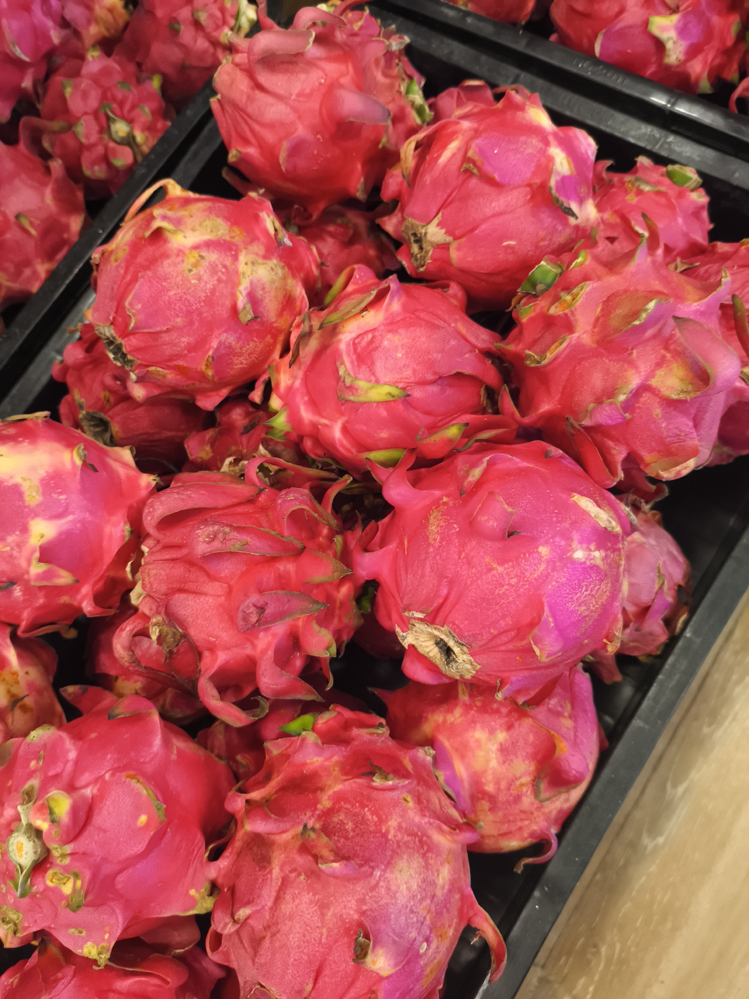 The image shows a close-up of a black plastic bin filled with red dragon fruit. The fruit are piled high, filling the container. The dragon fruit have a bright pink skin with green scales. The background is slightly out of focus, but it appears to be a market or grocery store setting. The fruits are close to being rotten or overripe. The scene is located in Depok, Indonesia.