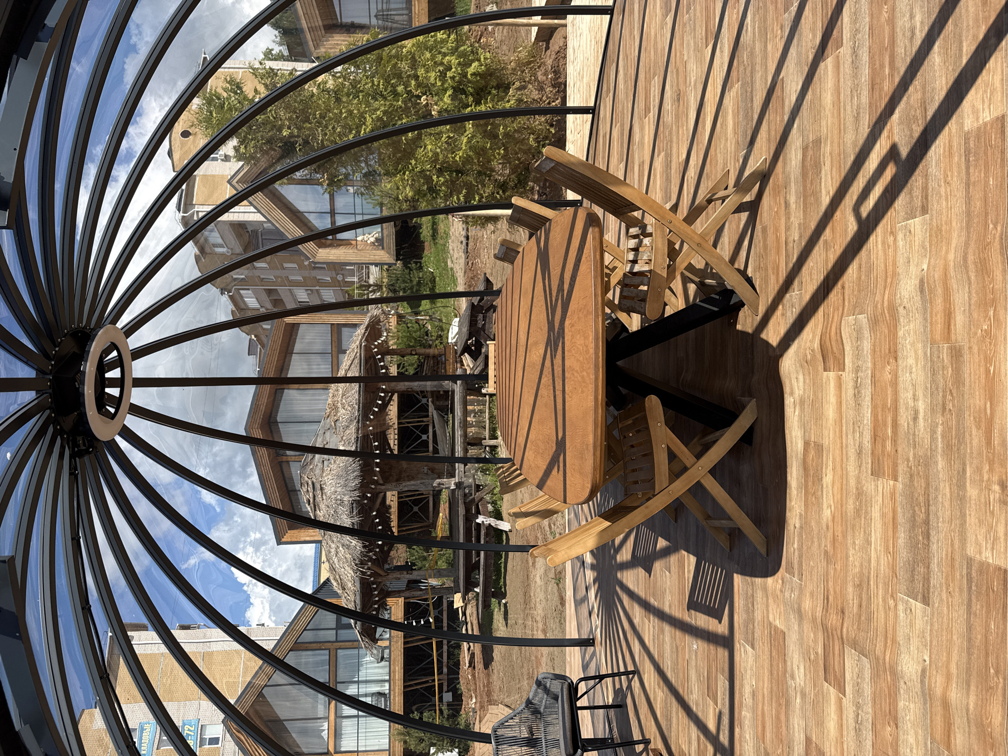 The image shows an outdoor patio setting, likely in the city of Votkinsk, Russia, based on the presence of structures with specific architectural characteristics common to the area. The focal point is a wooden table surrounded by wooden chairs, situated on a deck with wood-look flooring. A black metal framework, possibly part of a dome structure, arches overhead, offering views of surrounding buildings with unique rooflines. The area appears to be a public space with outdoor seating. Shadows cast by the structure and furniture create a dynamic interplay of light and dark across the flooring. The scene is daytime, under a partially cloudy sky visible through the dome's framework.
